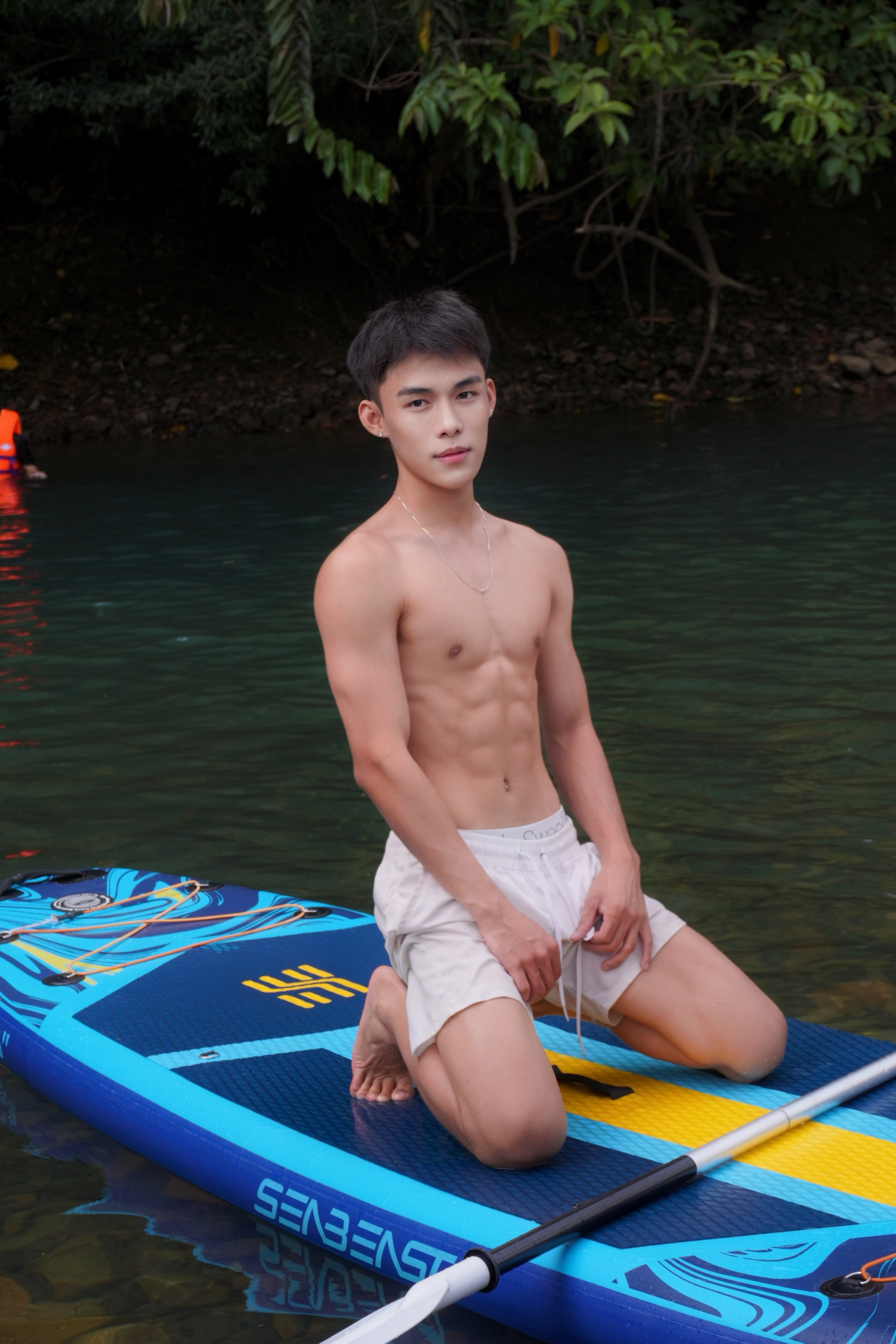 A young man with short dark hair and earrings, shirtless, kneeling on a blue stand-up paddleboard in a natural water setting, wearing cream-colored shorts and a silver necklace, with lush green foliage in the background.