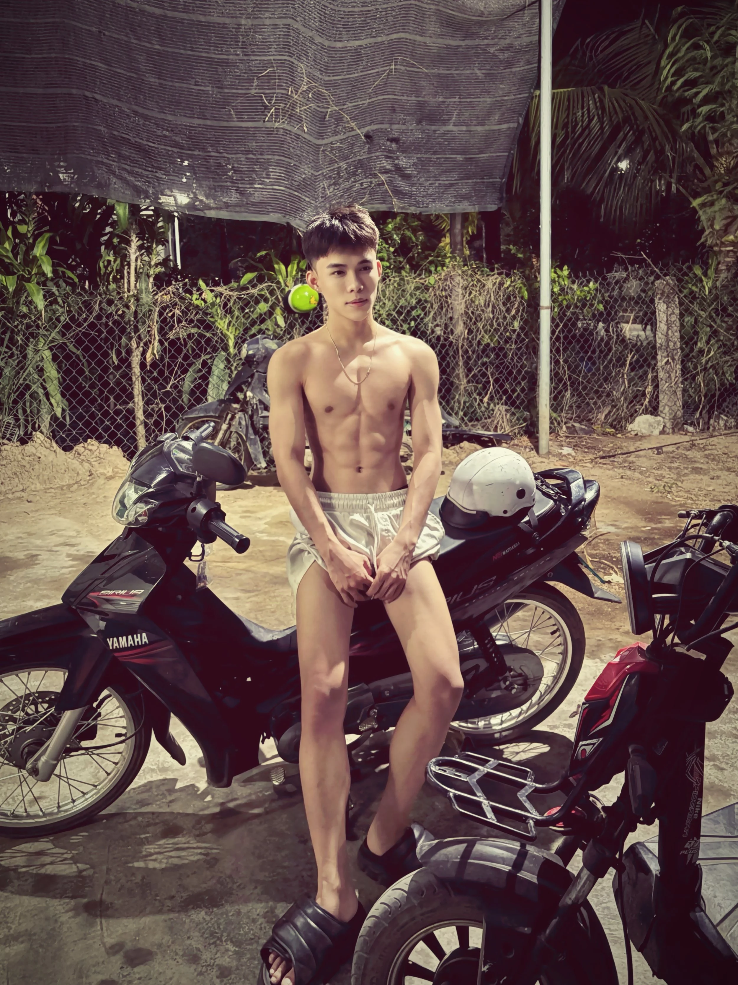 A young shirtless man with short hair standing next to a black Yamaha motorbike, surrounded by other motorbikes outdoors with a background of trees and a chain-link fence. He has a green ball hanging from his left ear, a helmet on the bike, and is wearing black slide sandals.