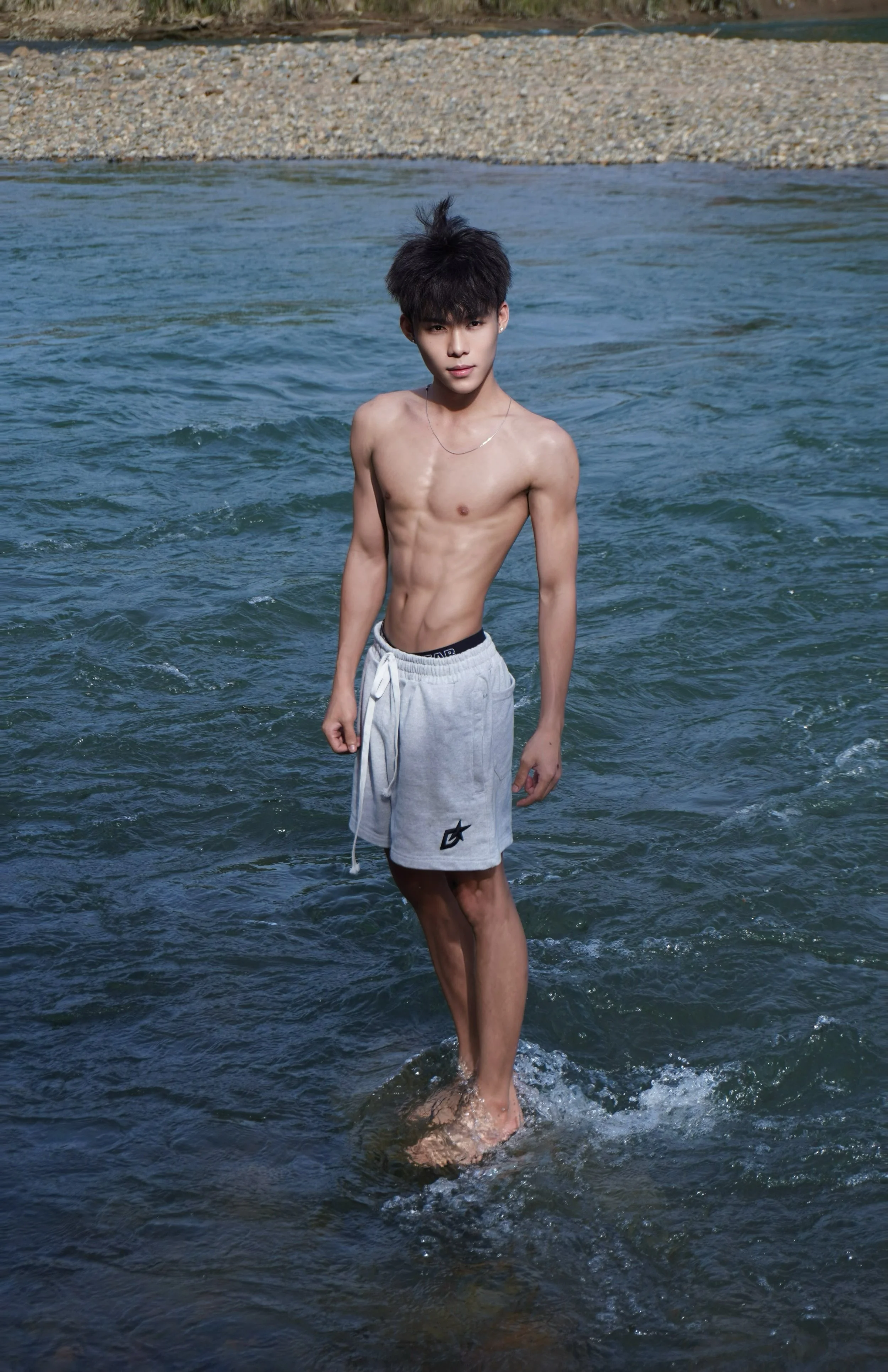 A shirtless young man with black hair in white gym shorts standing in a river with a stone-lined shore in the background.