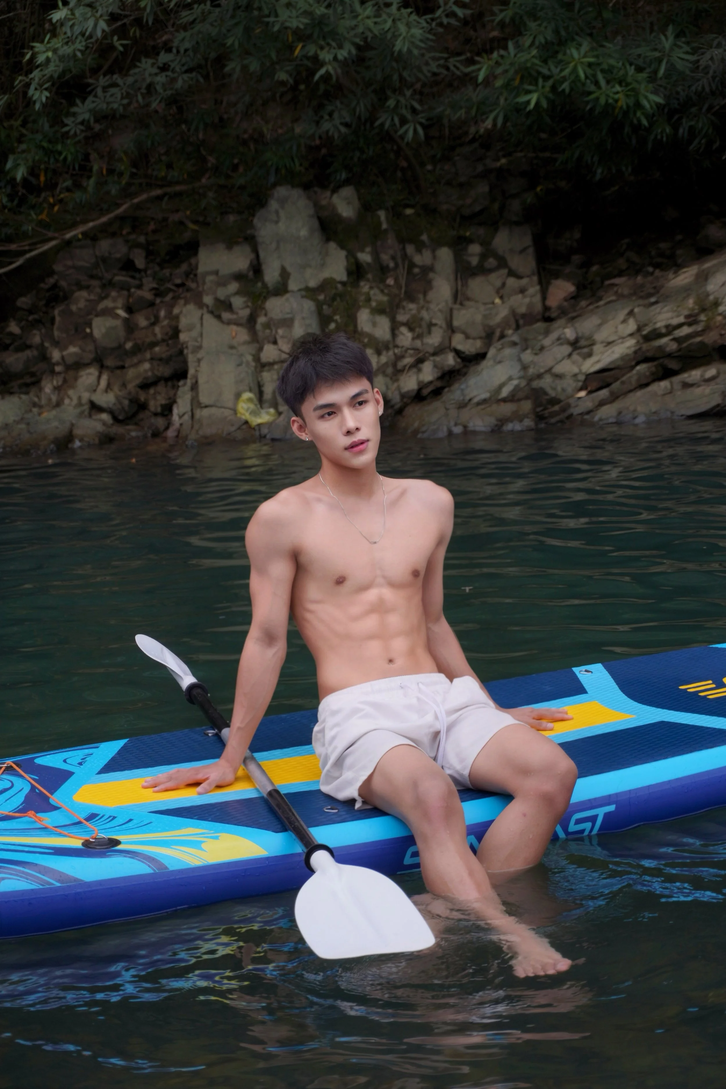 Young man with short dark hair and earrings, shirtless, sitting on an inflatable paddleboard in water, wearing khaki shorts, with a rocky and leafy background.