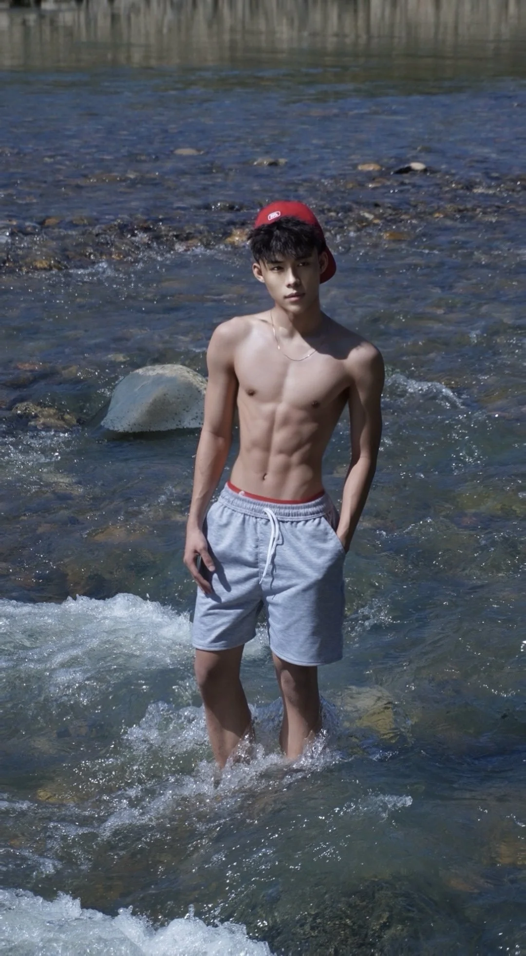A shirtless young man with black hair, wearing a red cap turned backwards and gray shorts, standing in a shallow rocky river.