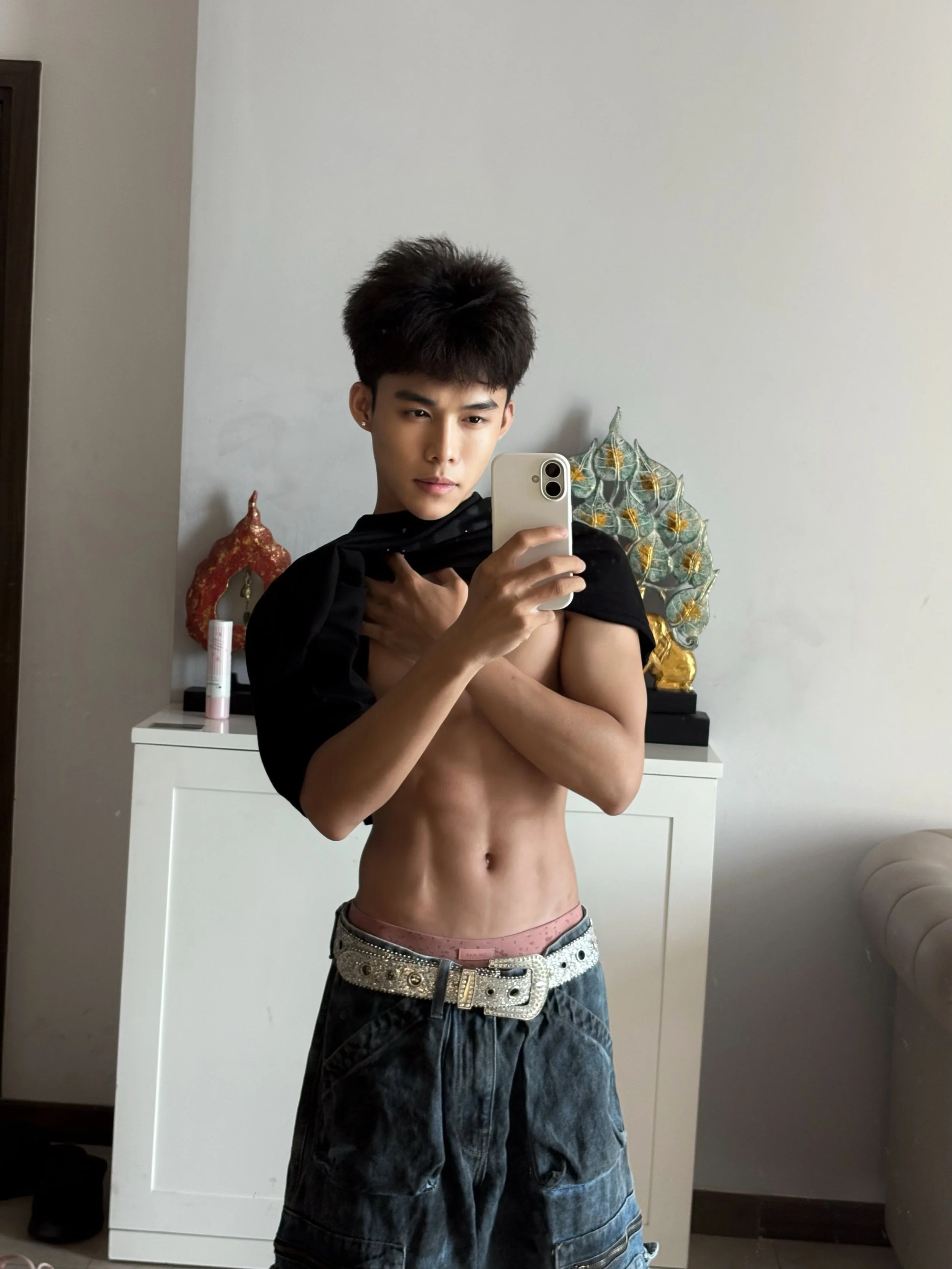 A young man is taking a mirror selfie, partially shirtless, wearing jeans with a decorative belt, and has a black cloth around his neck. There are decorative items on a white cabinet and art pieces on the wall behind him.