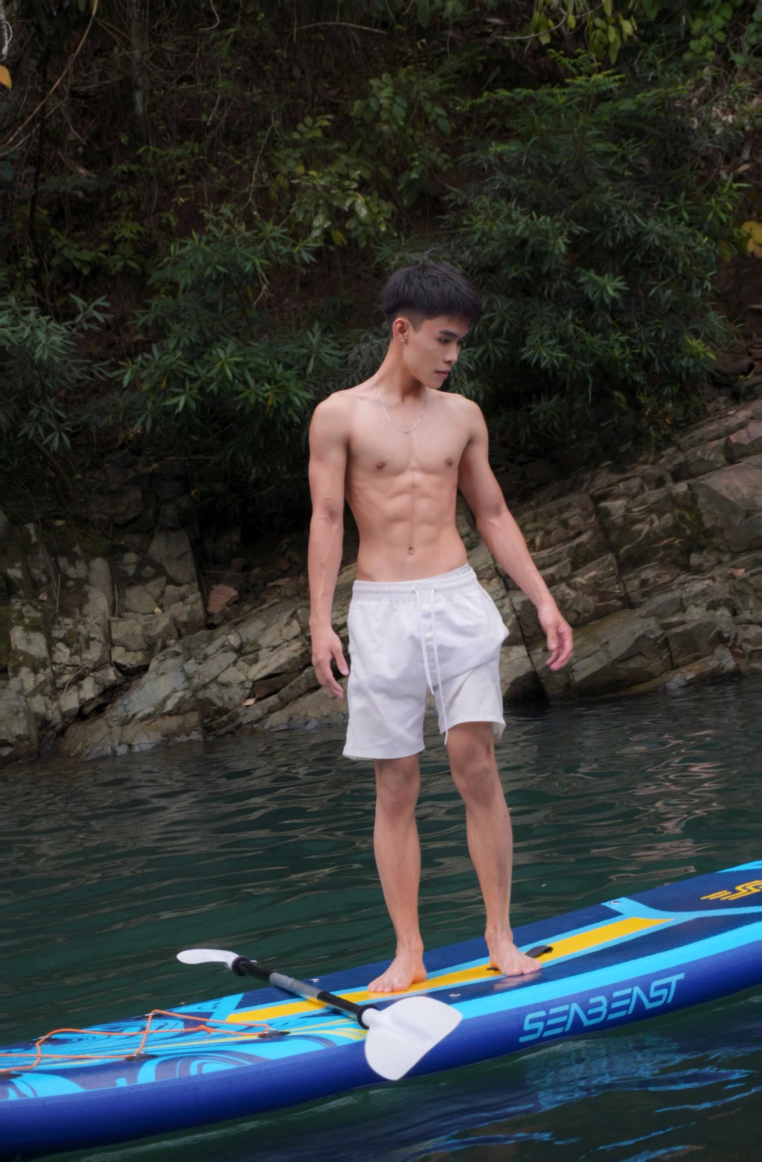 A young man with short dark hair, shirtless and wearing white shorts, stands on a blue stand-up paddleboard in a body of water. Behind him is a rocky shoreline with dense green foliage.