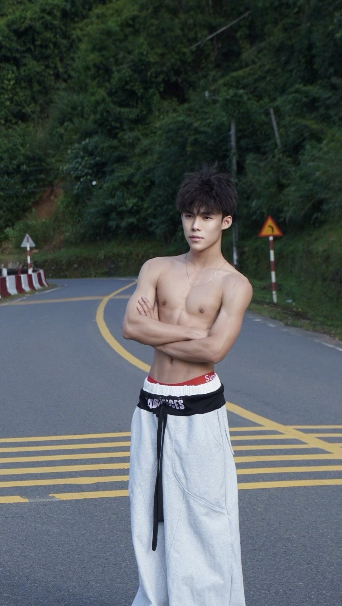 A shirtless young man standing on a winding road with arms crossed, wearing sweatpants with a red and white waistband, in a green forested area with road signs.