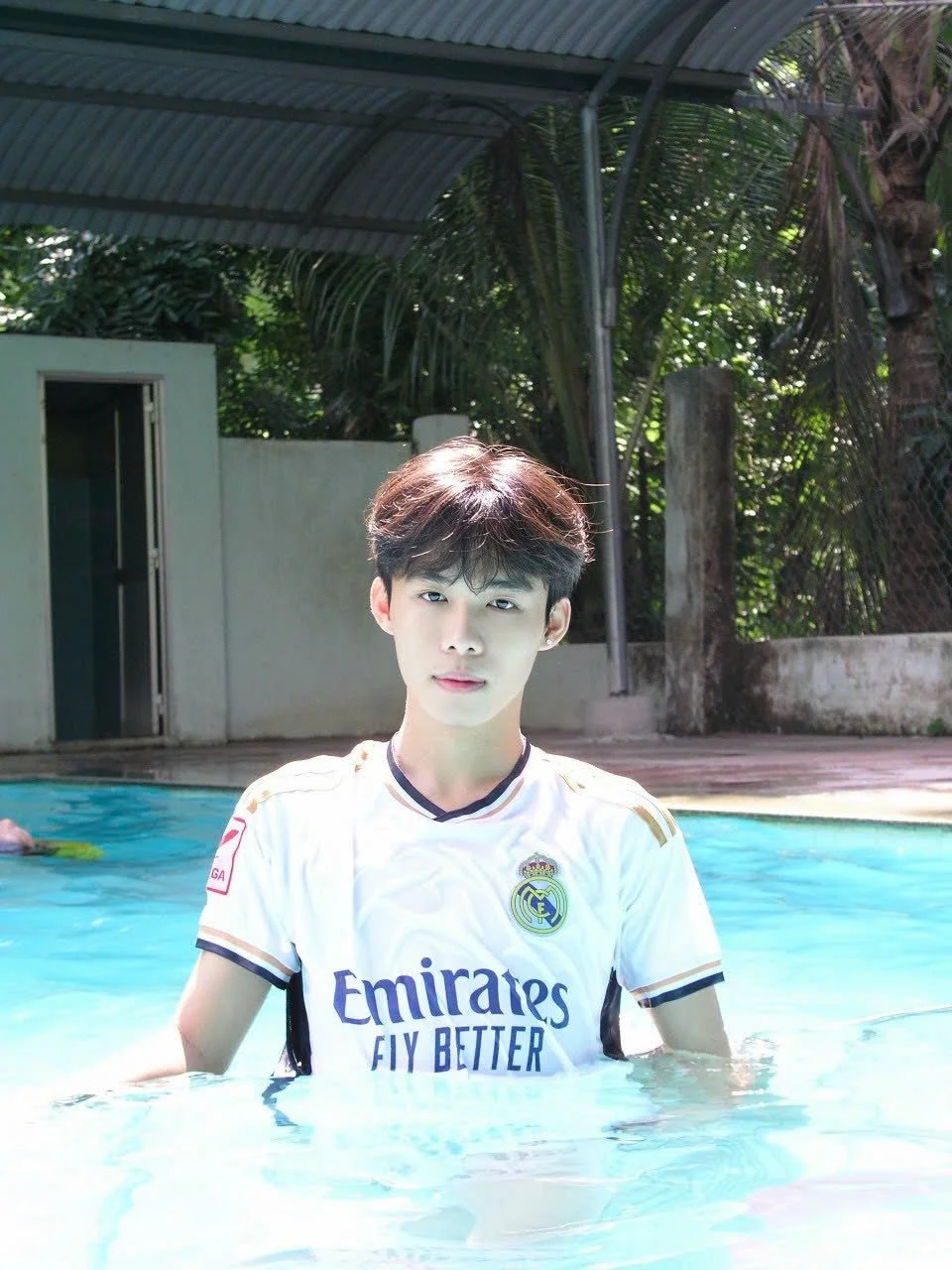 Young man in a white soccer jersey standing in a swimming pool during daytime, with a metal roof and green plants in the background.