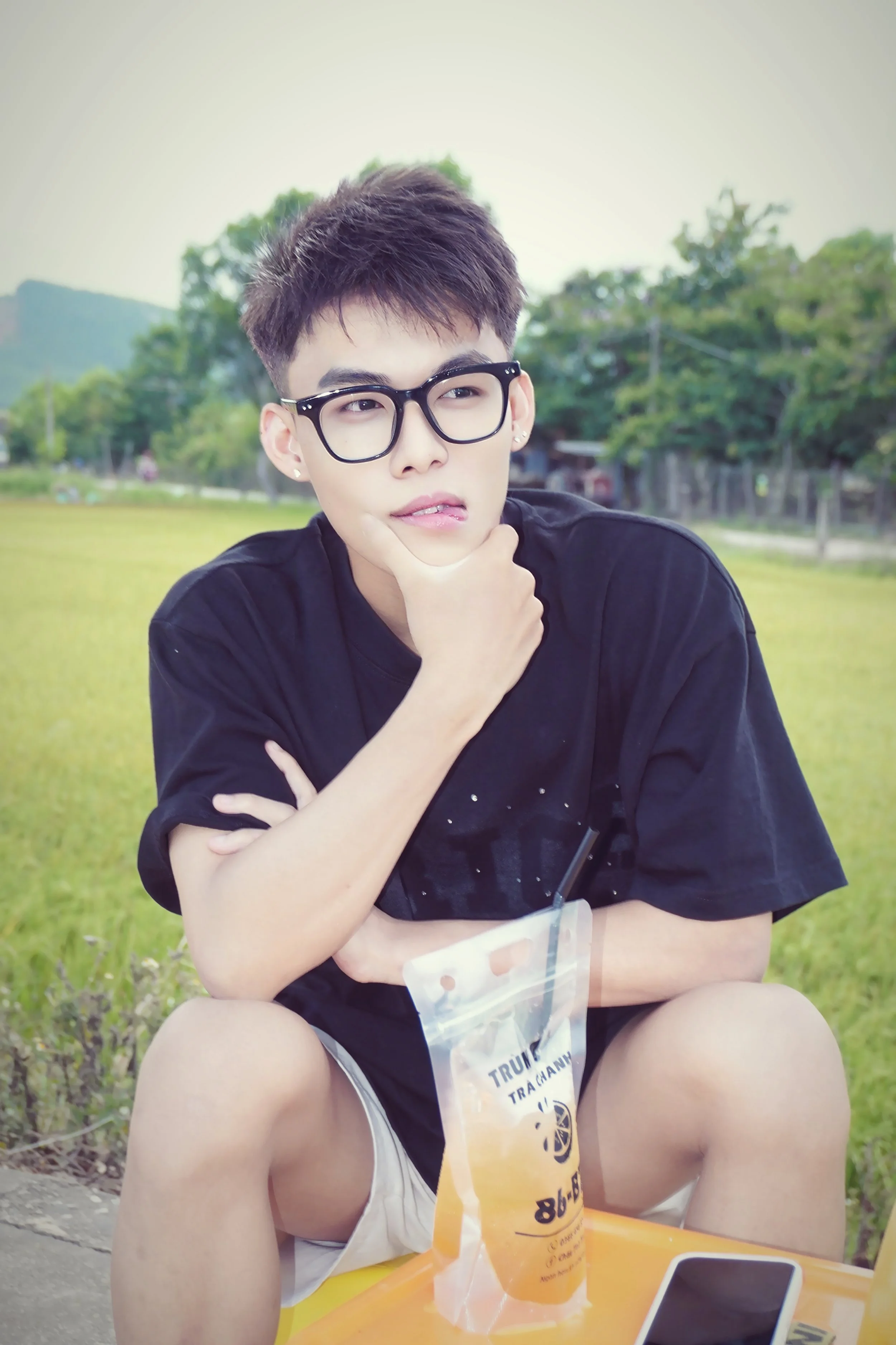 Young man with short dark hair, glasses, earrings, and a black T-shirt sitting outdoors at a yellow table with a drink in a plastic bag, a smartphone, and a tablet, in a grassy area with trees in the background.