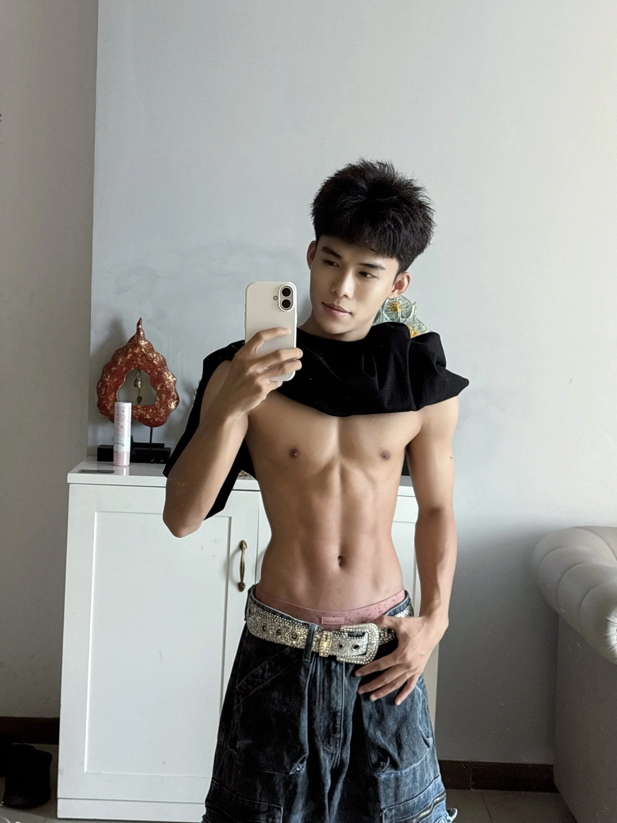 A young man taking a shirtless mirror selfie in a room with gray walls and white furniture. He is wearing jeans with a decorative belt and holding a smartphone. There is a red decorative object and some bottles on a white dresser behind him.