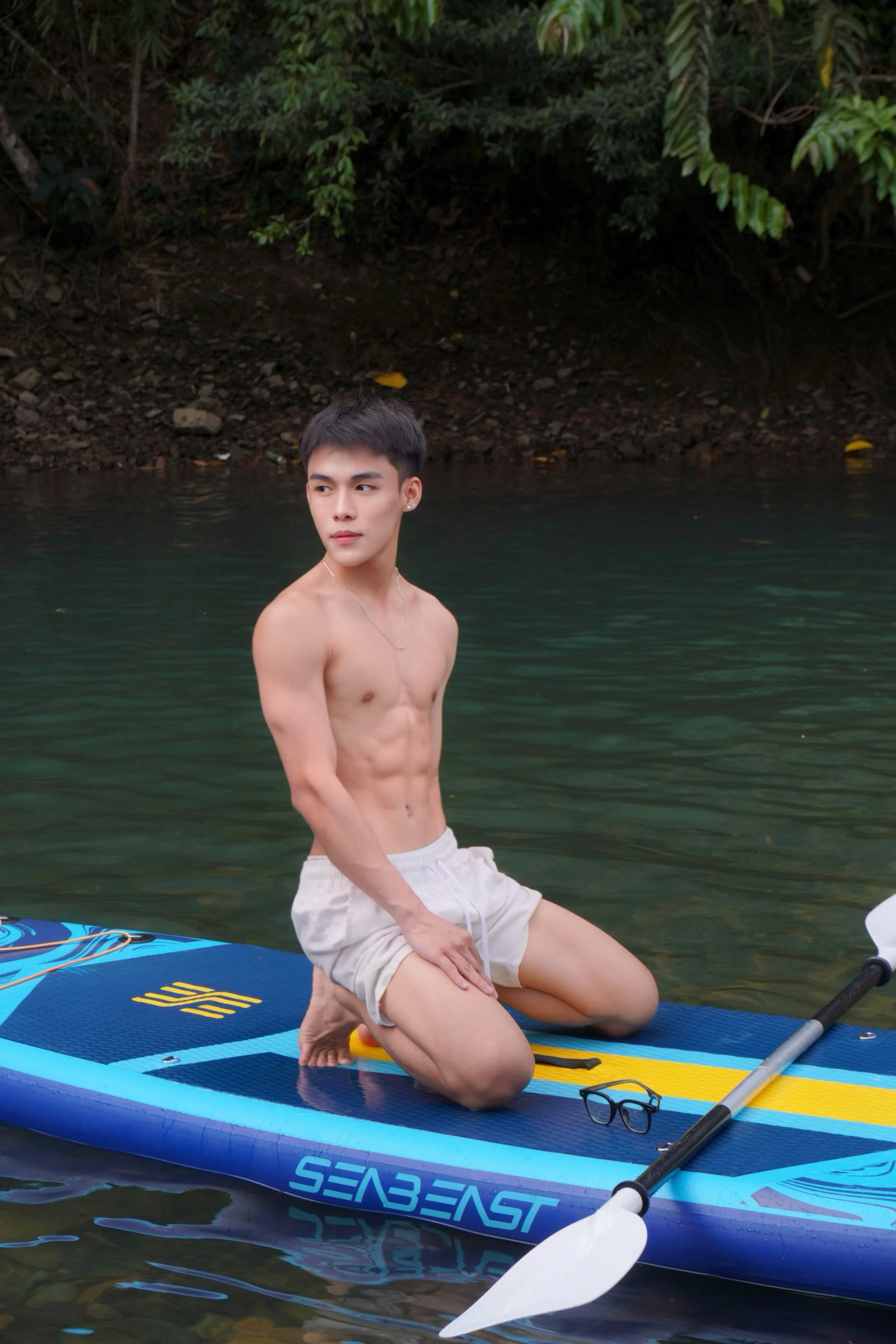A young man kneeling on a paddleboard in a river, surrounded by green trees, wearing beige shorts and no shirt, with sunglasses resting on the paddleboard.