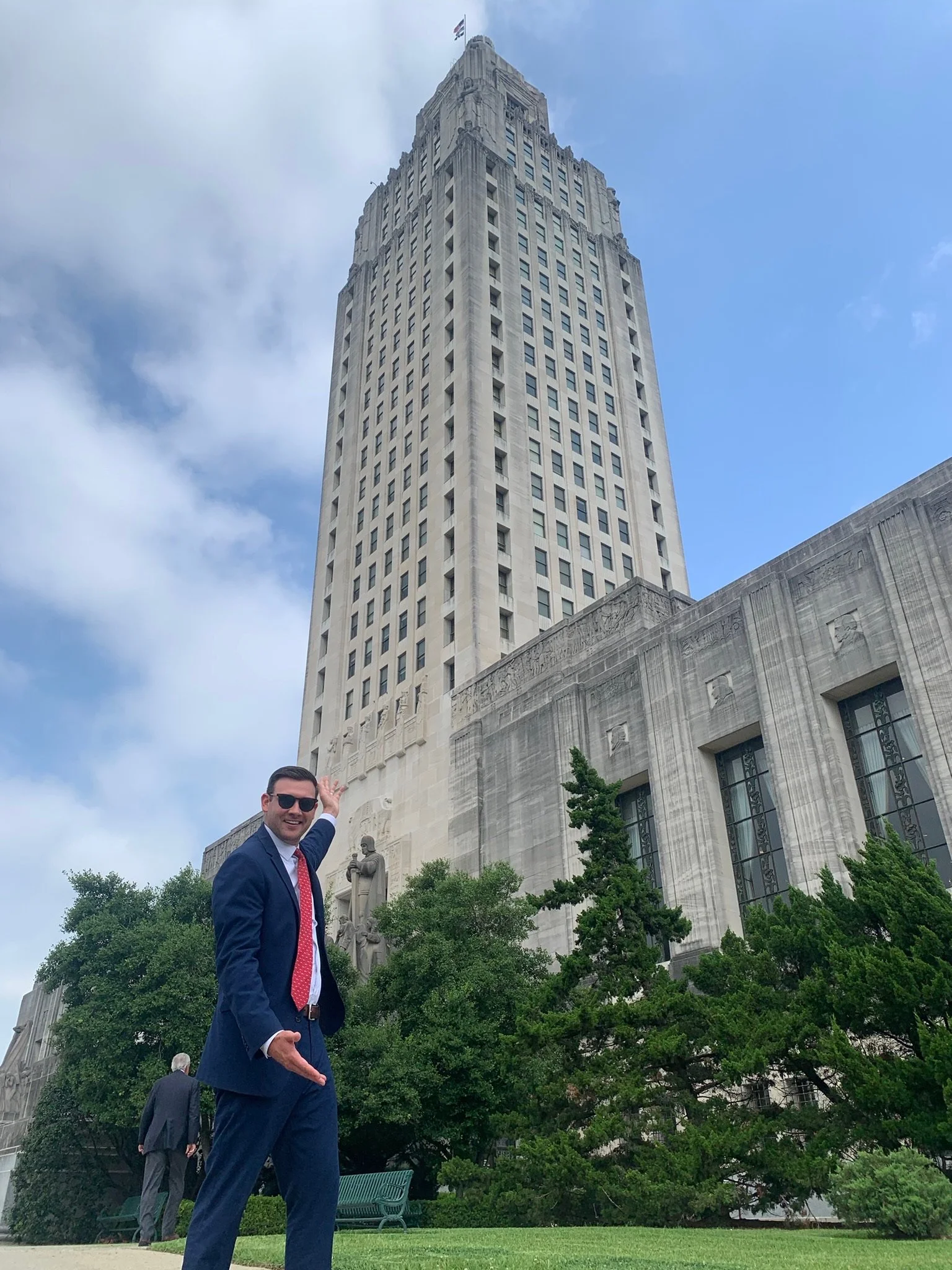 A man in a blue suit, red tie, and sunglasses points towards a tall, historic skyscraper with a flag on top, surrounded by trees and a park.