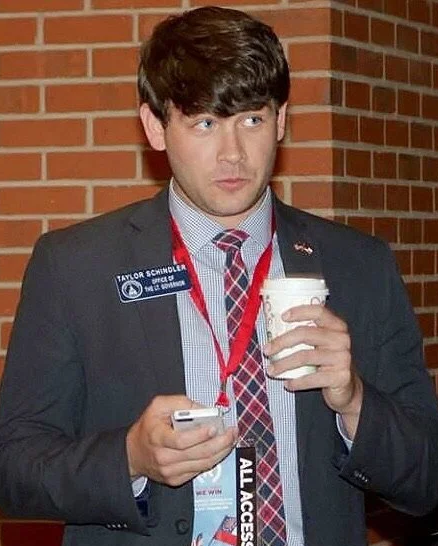 A young man in business attire, holding a coffee cup and a smartphone, standing in front of a brick wall. He has a name badge and an event lanyard around his neck.