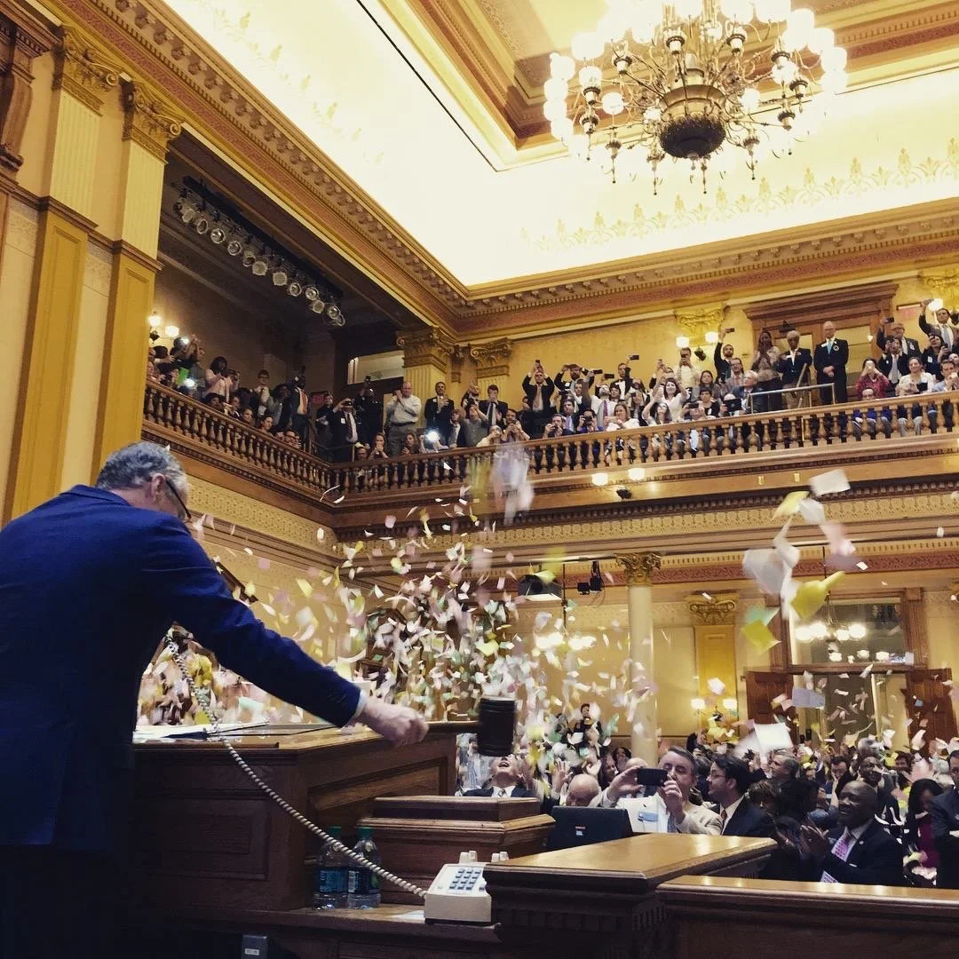 A large, ornate room with a crowd gathered, many taking photos or recording as confetti falls. A person in a suit at a podium is throwing confetti among applauding attendees, some seated and others on a balcony overlooking the scene. The room has gold accents and a large chandelier.