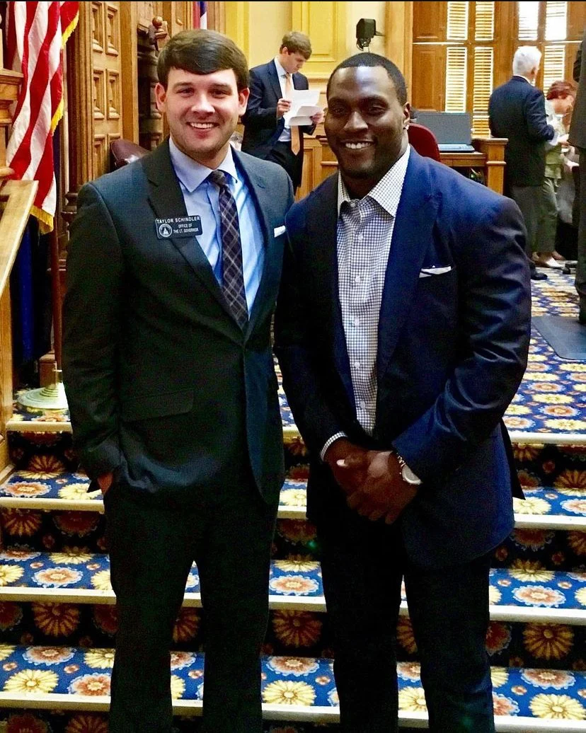 Two men in suits standing side by side in a formal room with a colorful patterned carpet, wooden paneling, and other people in the background engaged in conversation or reading. The man on the left is smiling, wearing a name tag that says 'Taylor Sch