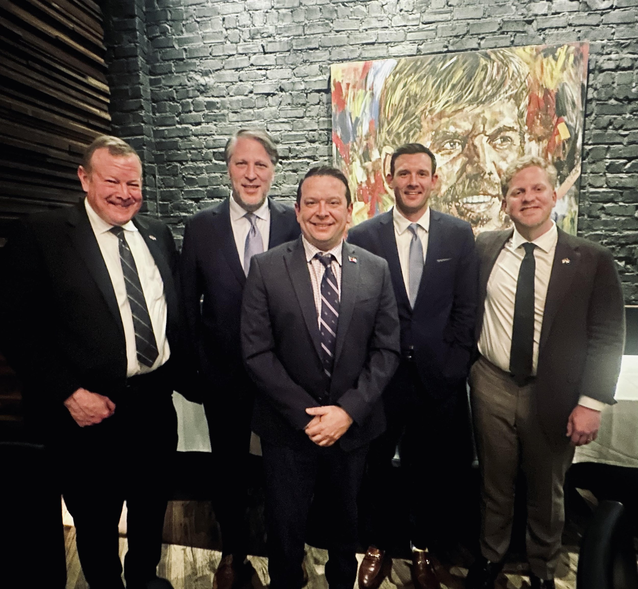 Group of five men in suits standing together in front of a black brick wall and a colorful portrait of a man with glasses.