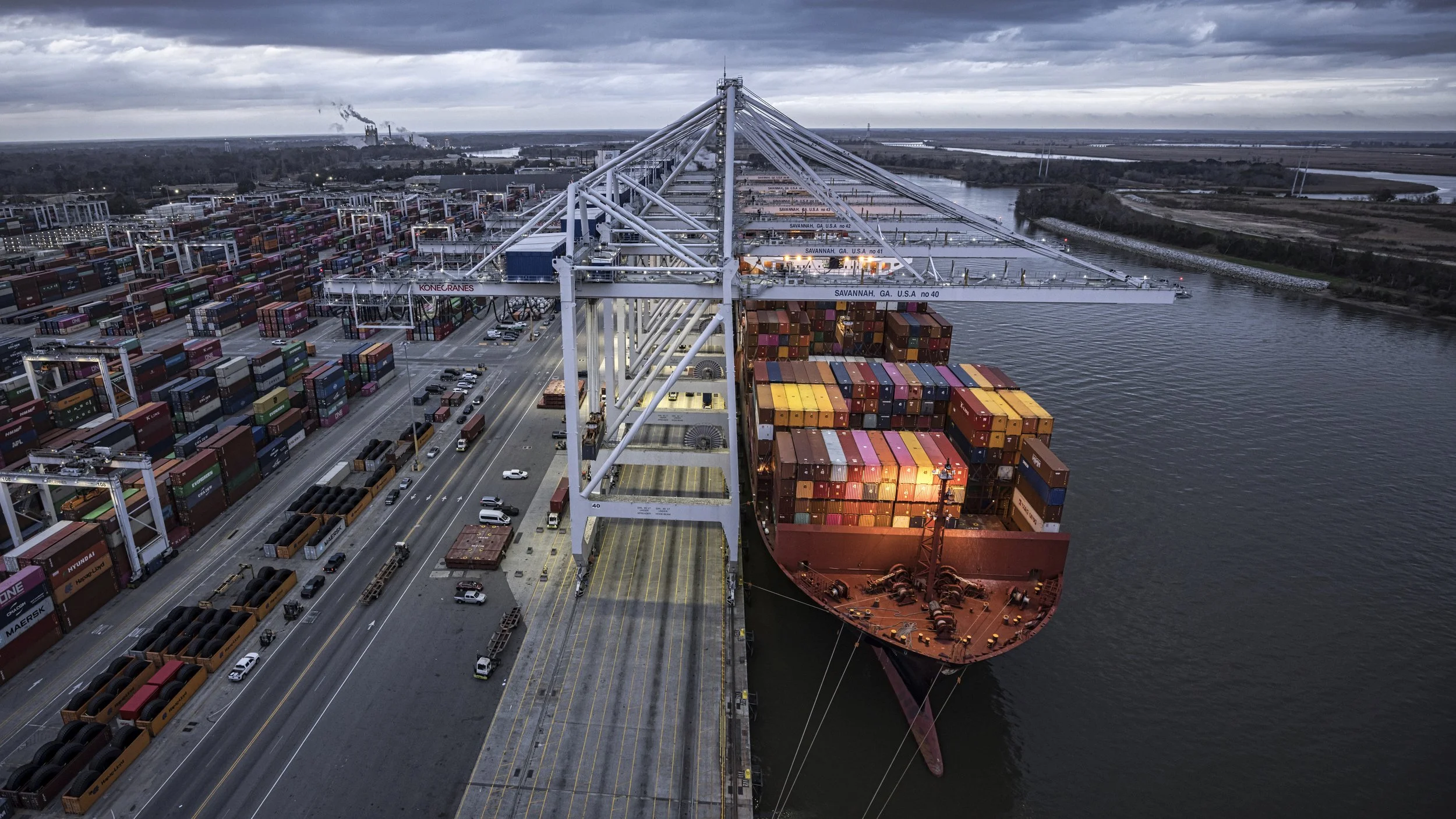Aerial view of a busy container port with cargo ships docked, surrounded by stacked containers, and large cranes for loading and unloading, with industrial buildings and waterways in the background.