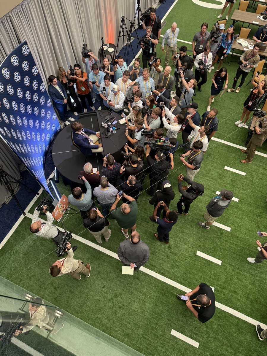 A man in a suit sitting at a round table at a press conference surrounded by numerous reporters, photographers, and videographers capturing the event, with a SEC banner on the left and a press room setting.