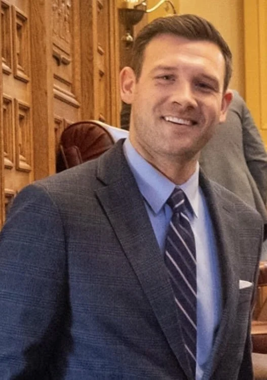Man in a suit smiling in a formal setting with wooden paneling in the background.