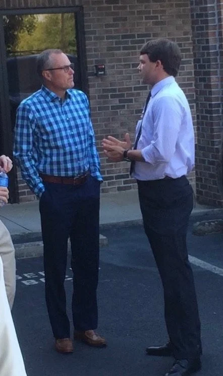 Two men are having a conversation outside a brick building. One man is wearing a blue checkered shirt and glasses and has his hands in his pocket. The other man is wearing a white shirt with a tie and is gesturing with his hands.