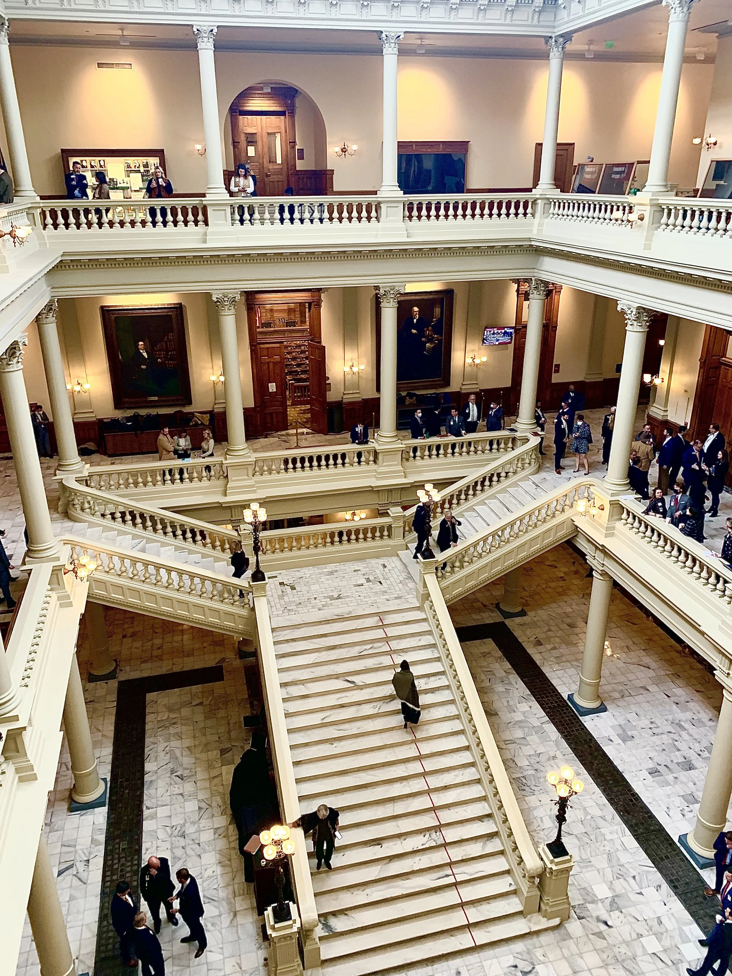 Elegant, multi-story interior of a historic building with a grand staircase, marble floors, decorative columns, and people in formal attire walking and socializing.