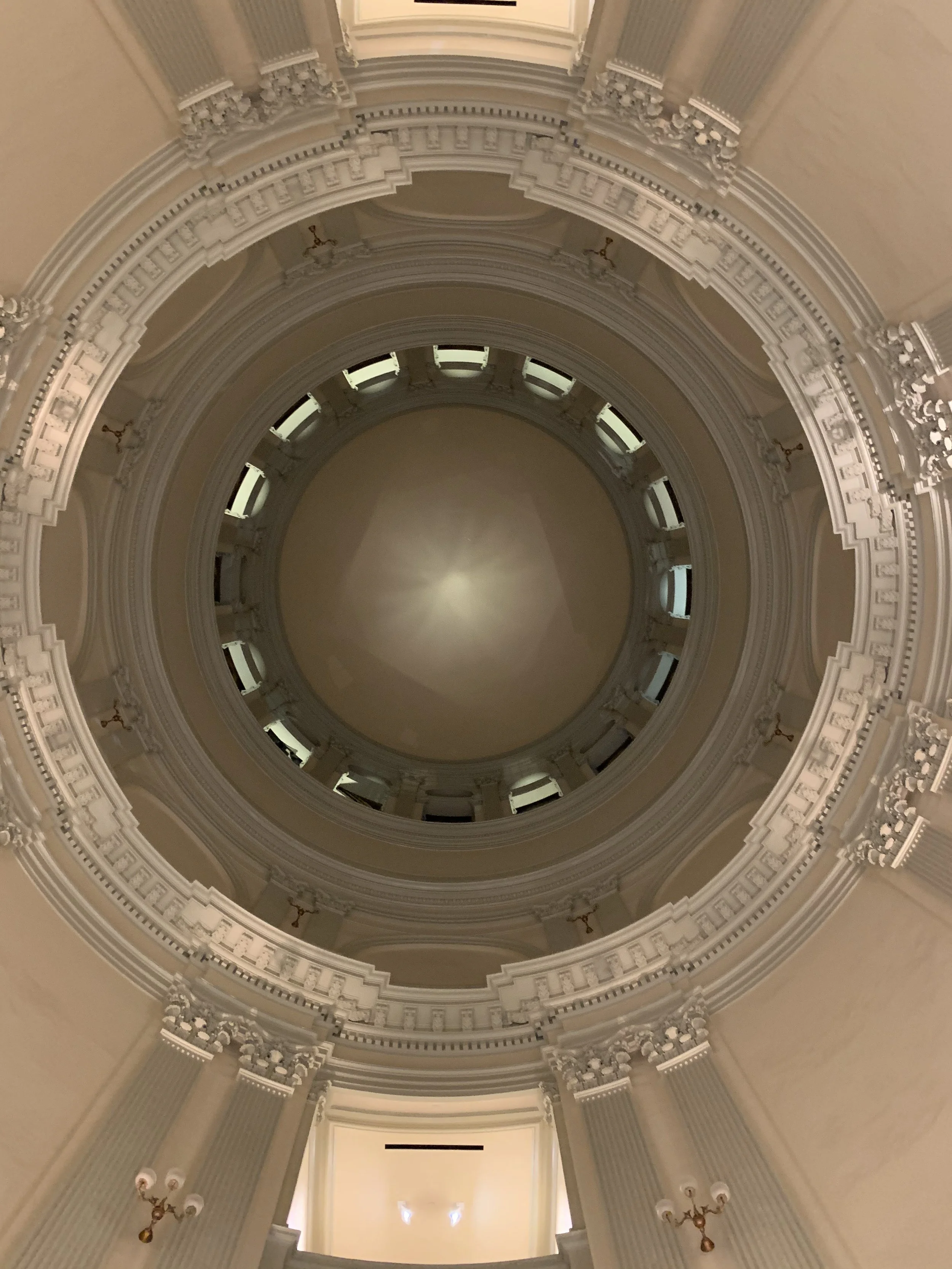 Looking up at a tall, ornate, circular interior ceiling with multiple layers of intricate molding and decorative elements, including small windows around the upper ring.