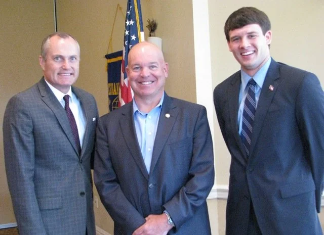 Three men in suits standing in front of a U.S. flag, smiling for the photo.