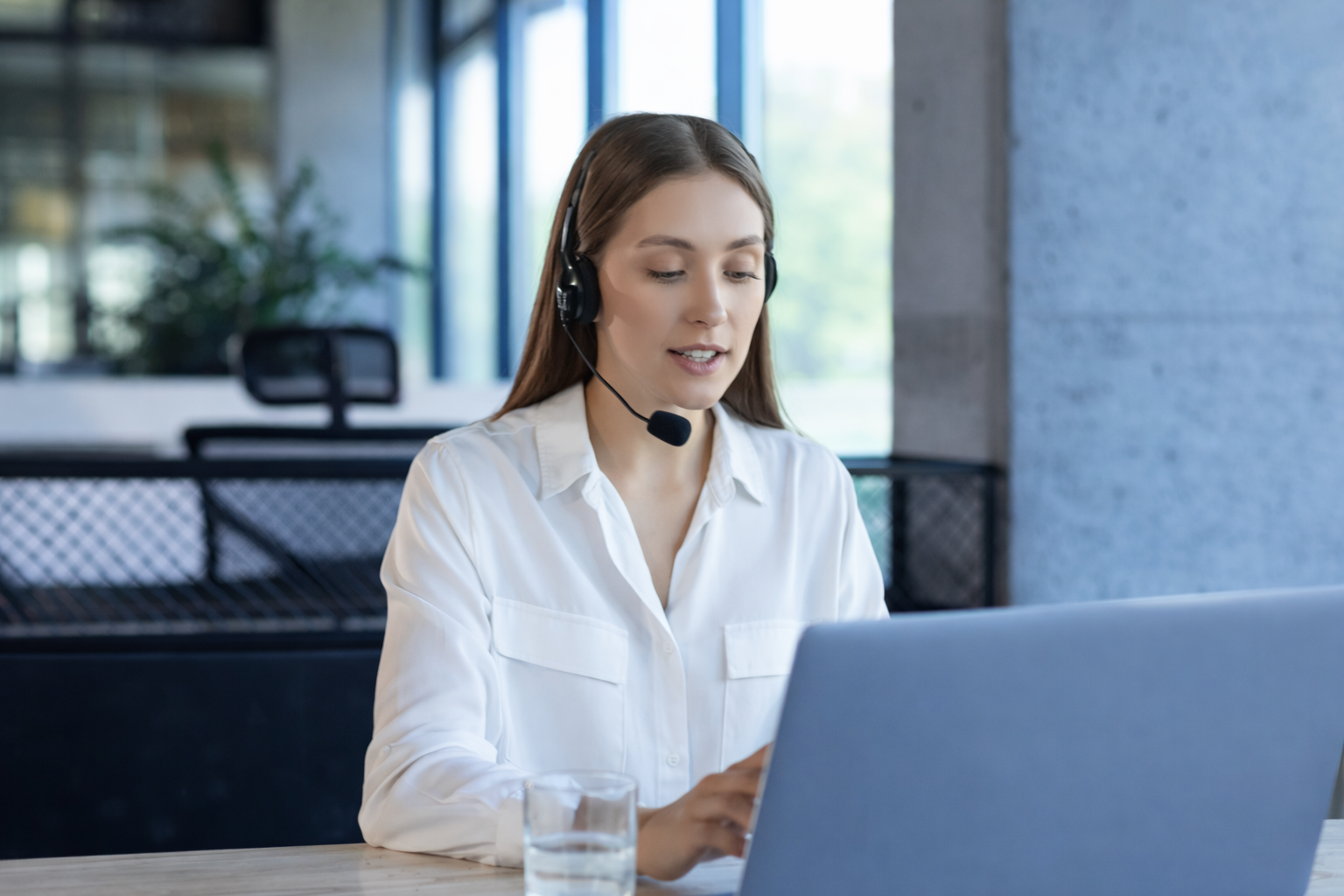 Mujer con auriculares de diadema y micrófono trabajando en una computadora portátil en una oficina moderna.