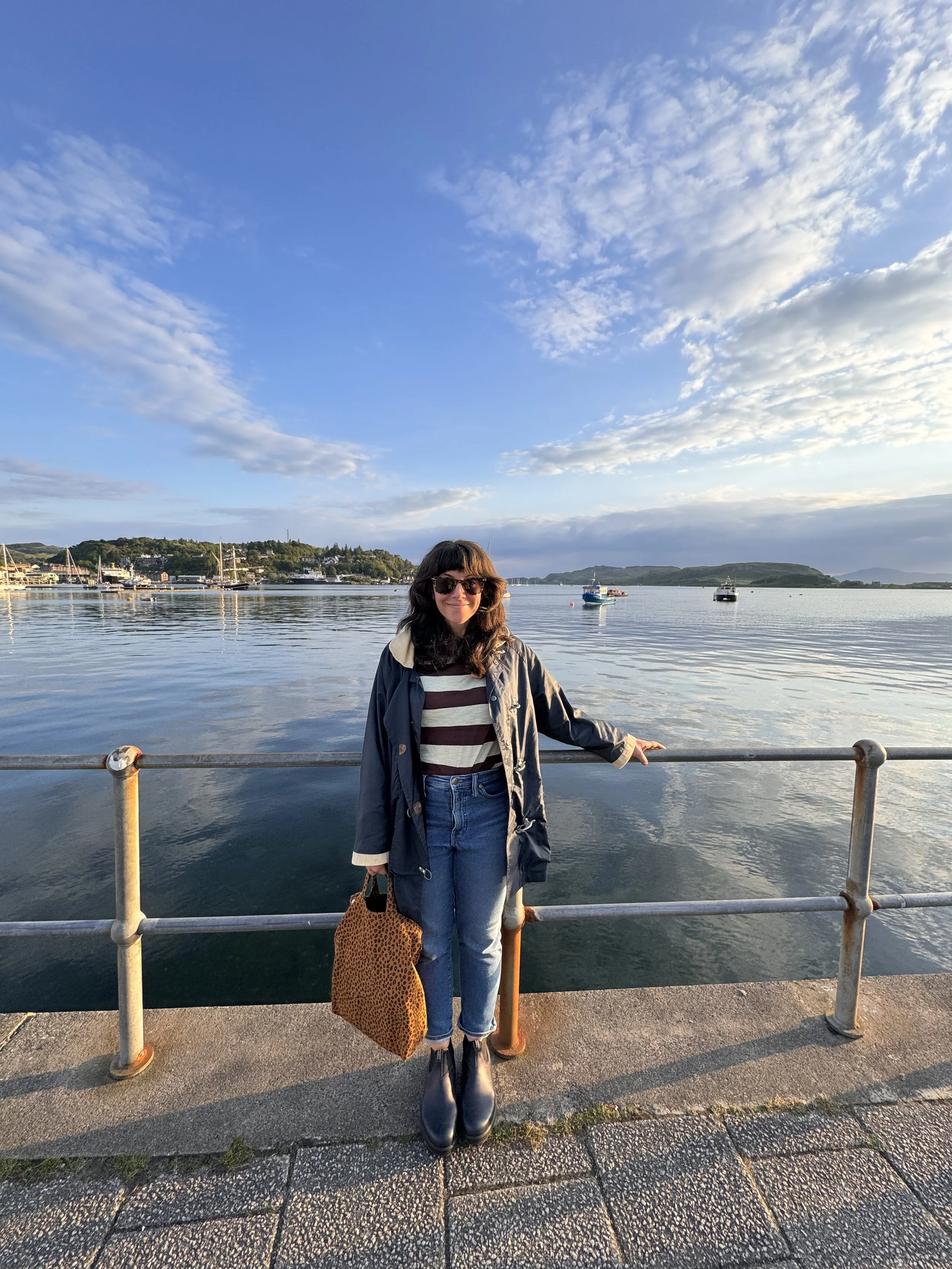 Mary Karmelek- woman standing by a waterfront railing during sunset, with water and boats in the background, wearing sunglasses, a striped shirt, a jacket, jeans, and holding a leopard print bag.