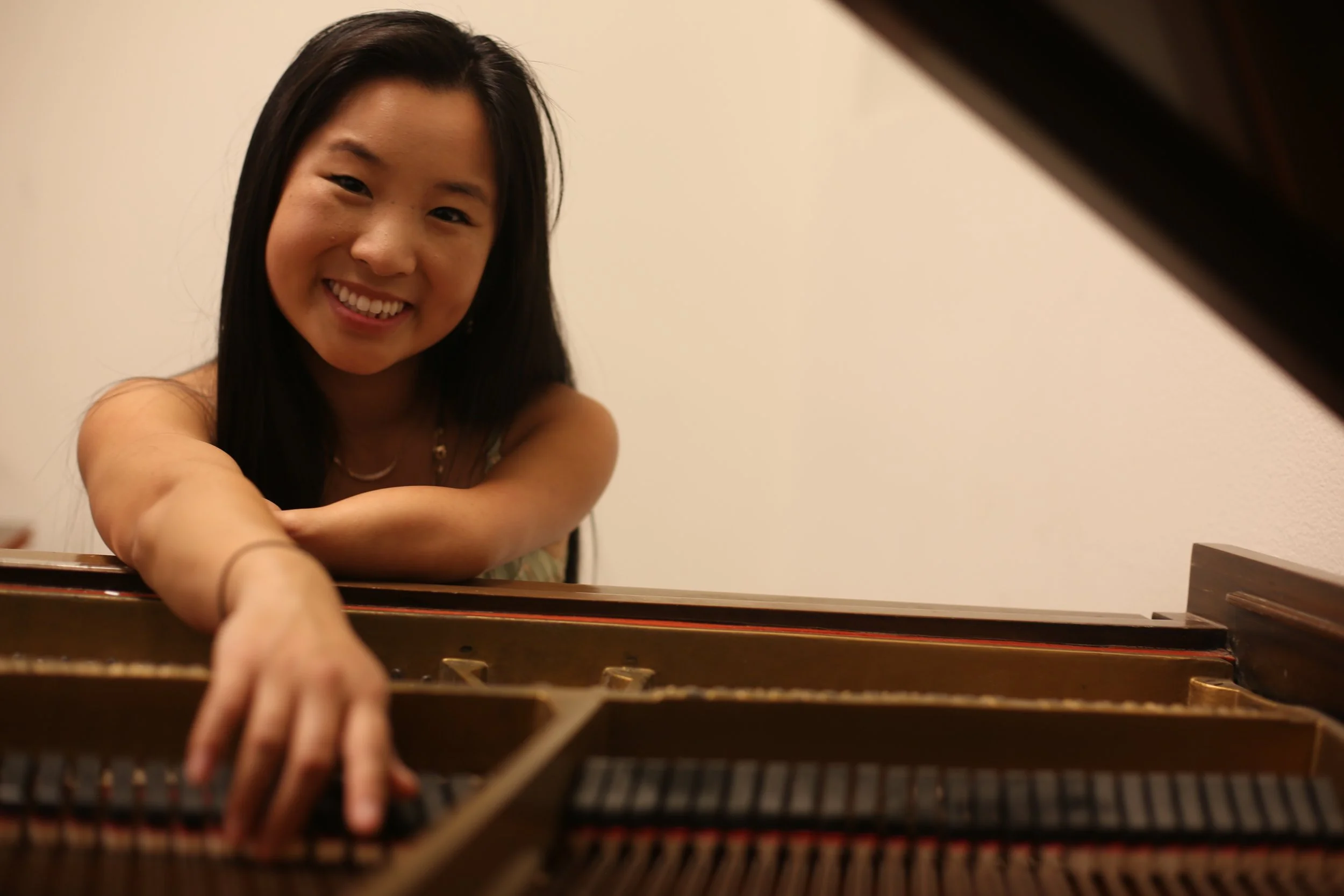 A smiling woman with long black hair reaches towards the camera over a piano, with a plain beige wall in the background.