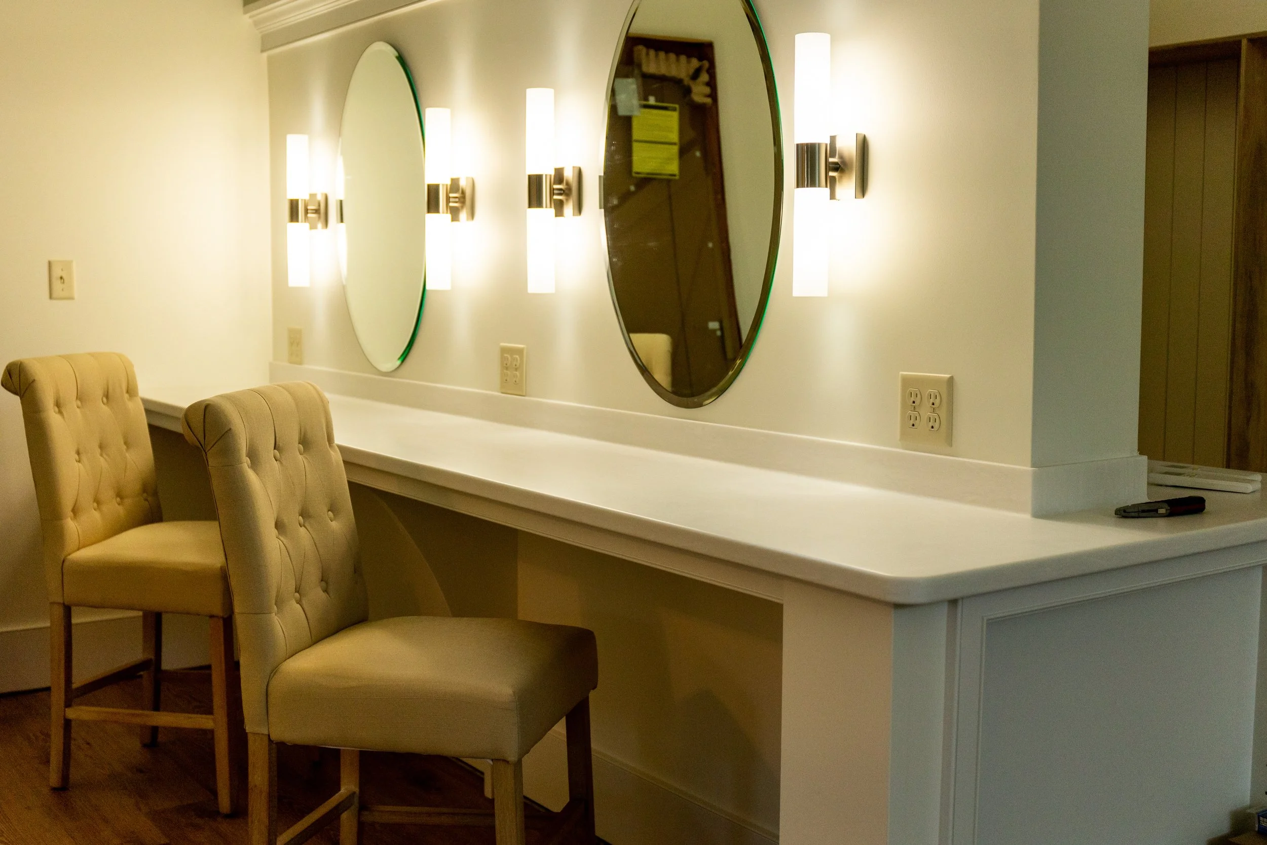 A hotel or salon vanity area with two beige tufted chairs, a long white countertop, two large oval mirrors, and wall-mounted light fixtures.