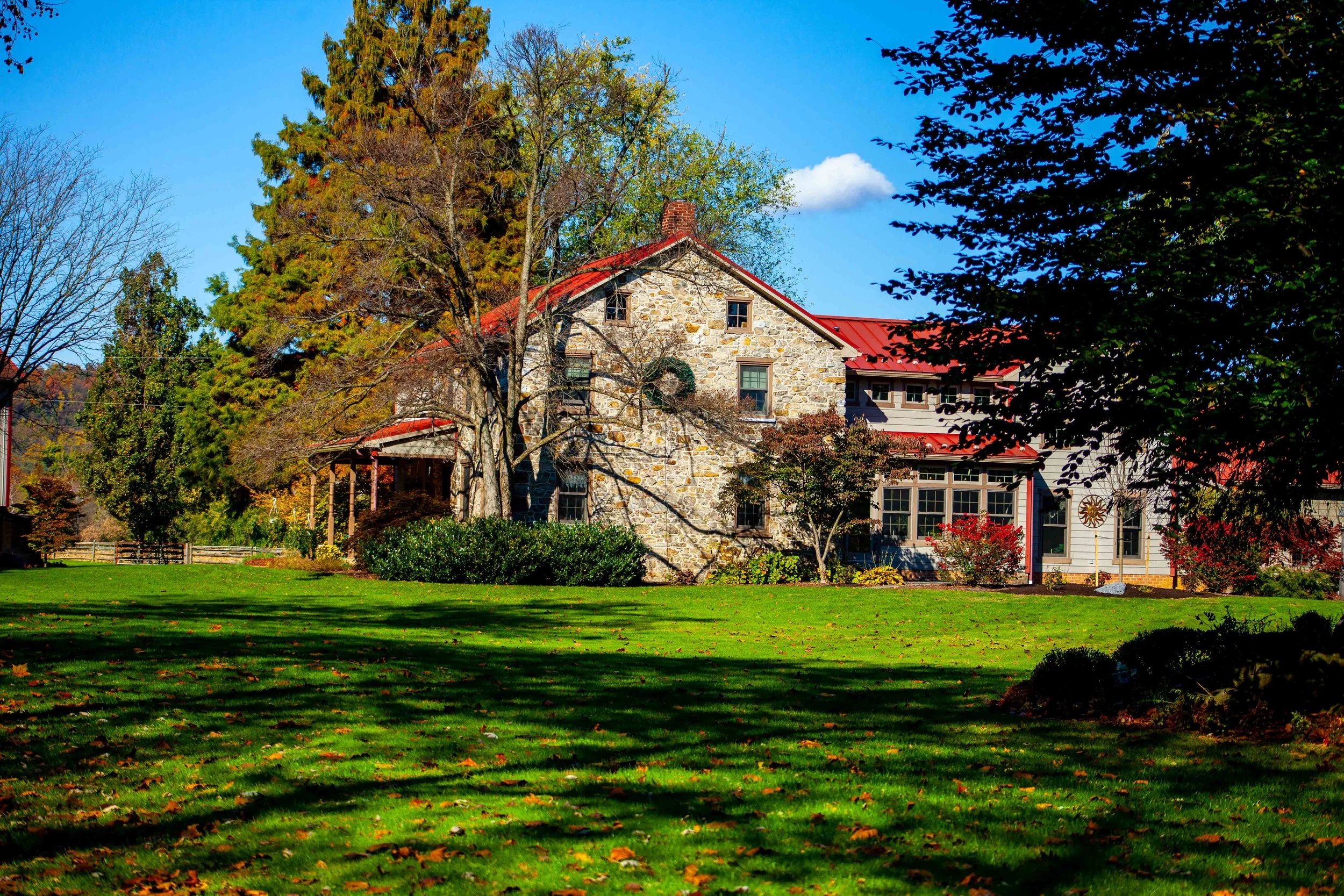 A large stone house with a red metal roof surrounded by trees with fall foliage, green lawn with shadows, and a blue sky with clouds.