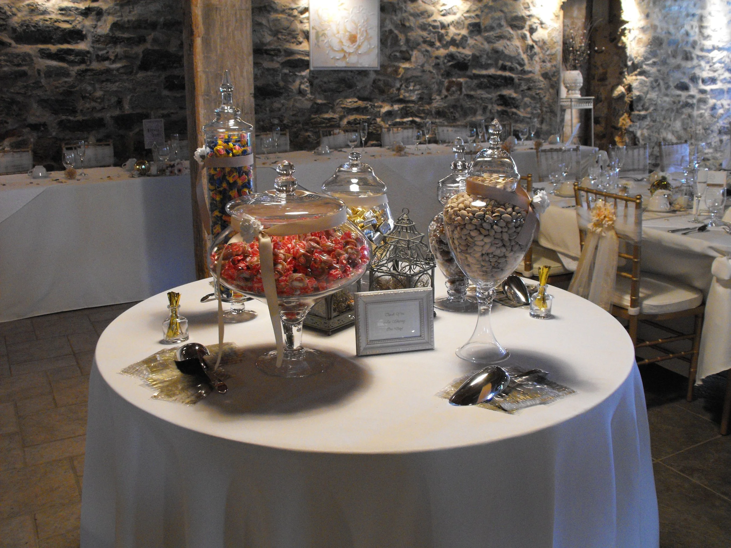 Round table with a white tablecloth displaying candy in glass jars at a wedding reception, with chairs and more tables in the background against stone walls.