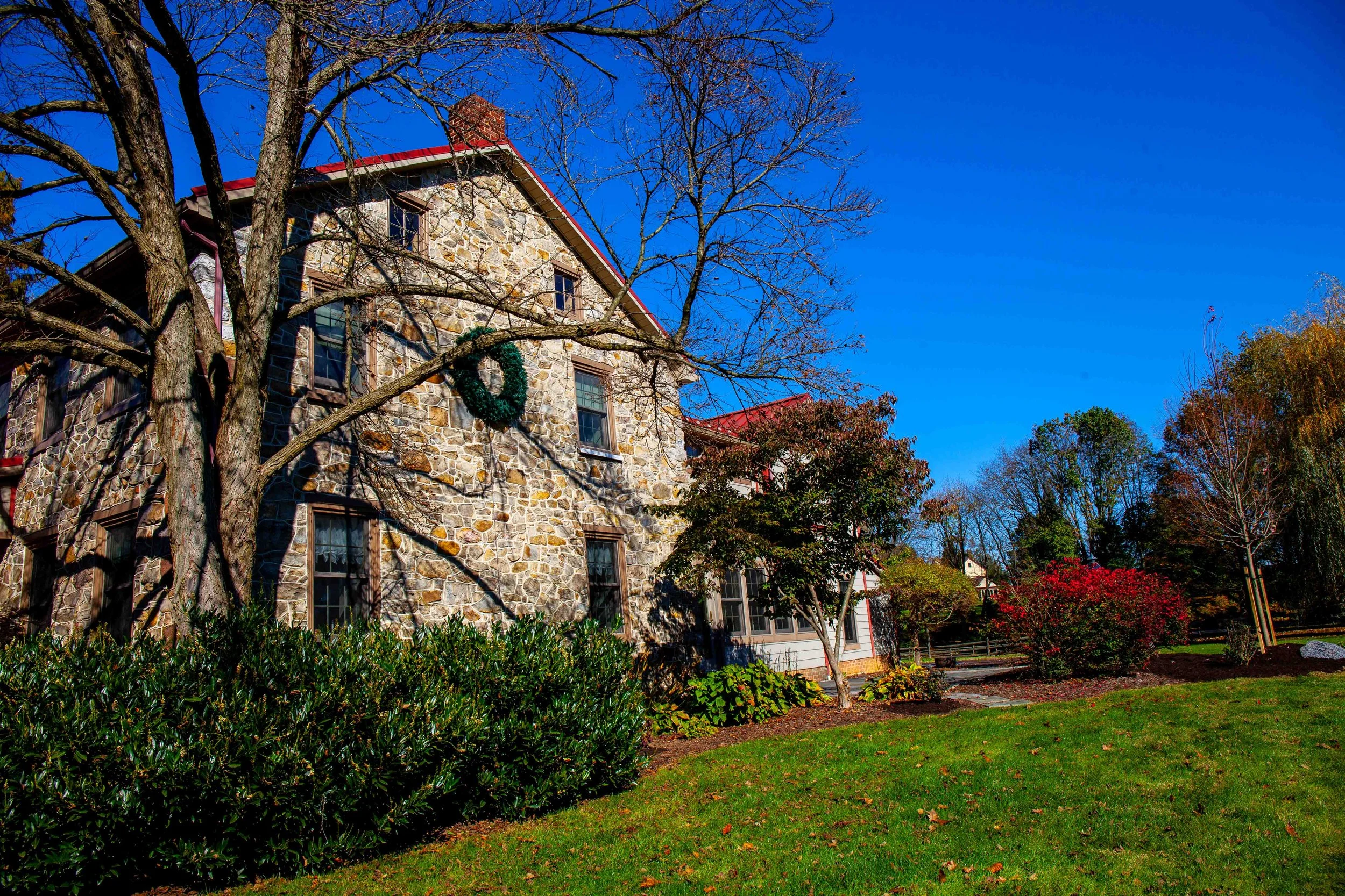 A stone house with red roof and multiple windows, surrounded by trees and bushes with fall foliage, under a clear blue sky.