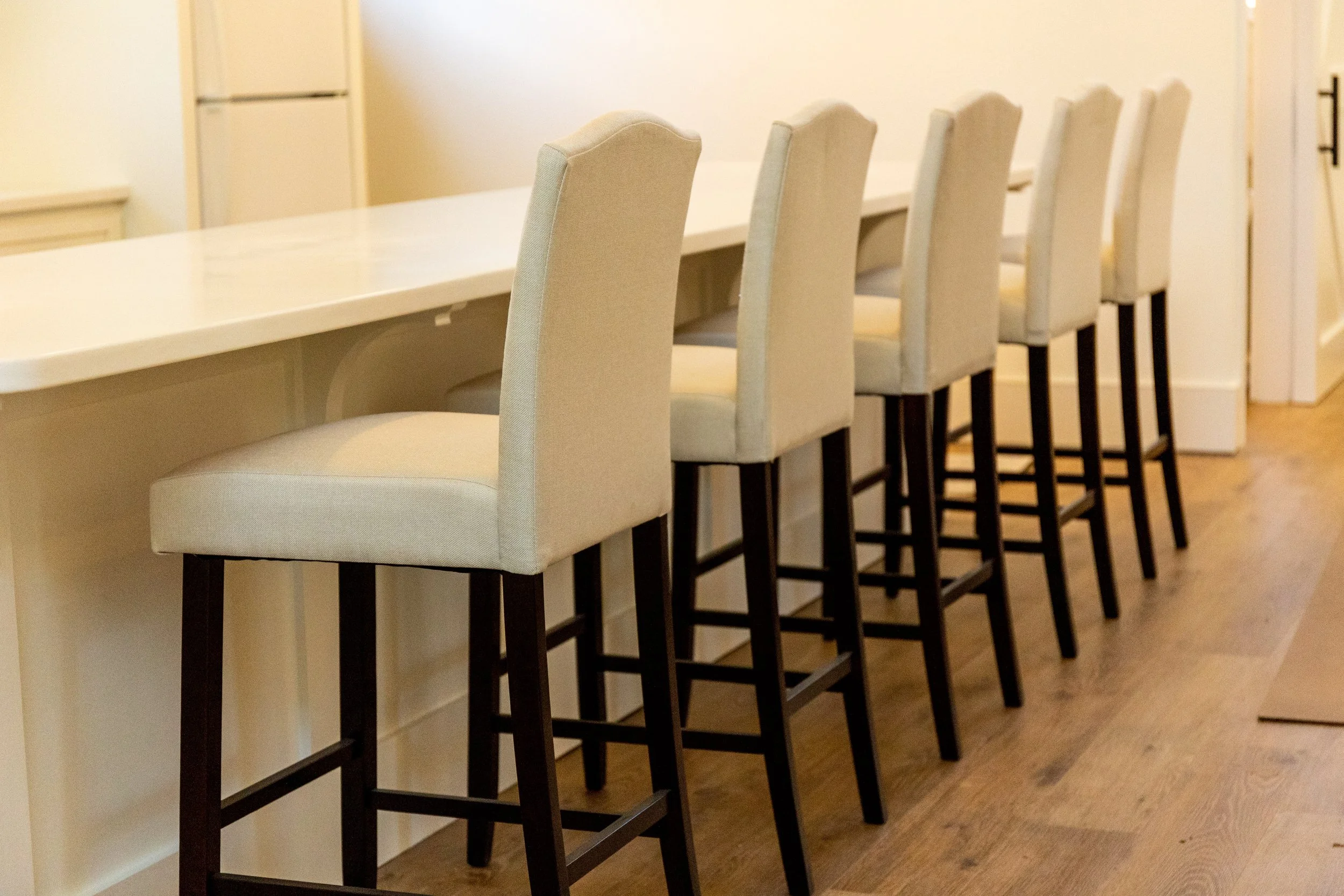 A row of six beige upholstered bar stools with dark wooden legs along a white kitchen island.