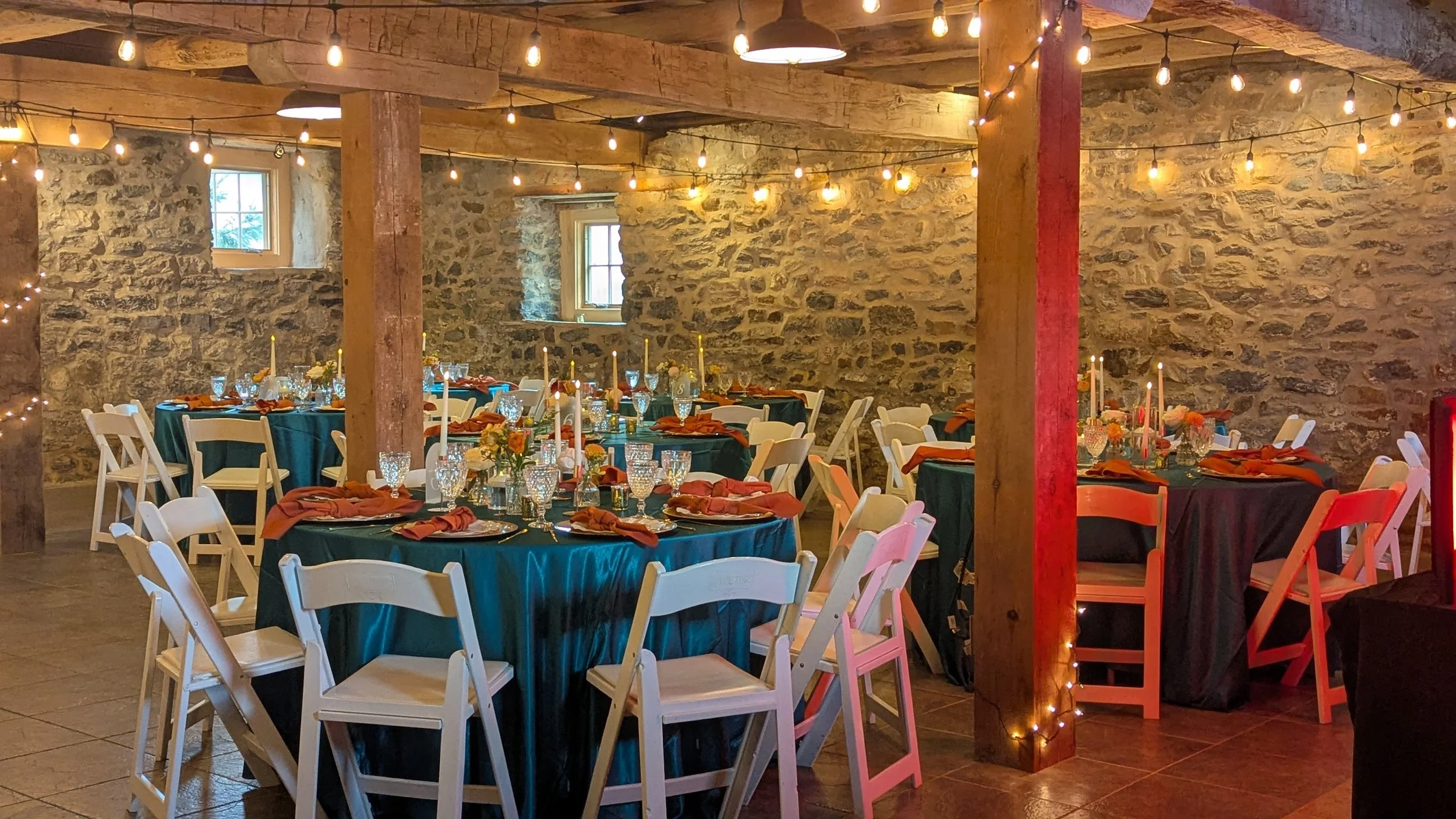 Radiant wedding or event banquet hall decorated with string lights, rustic stone walls, and round tables covered with dark tablecloths, set with glassware, candles, and floral centerpieces, with white chairs arranged around the tables.