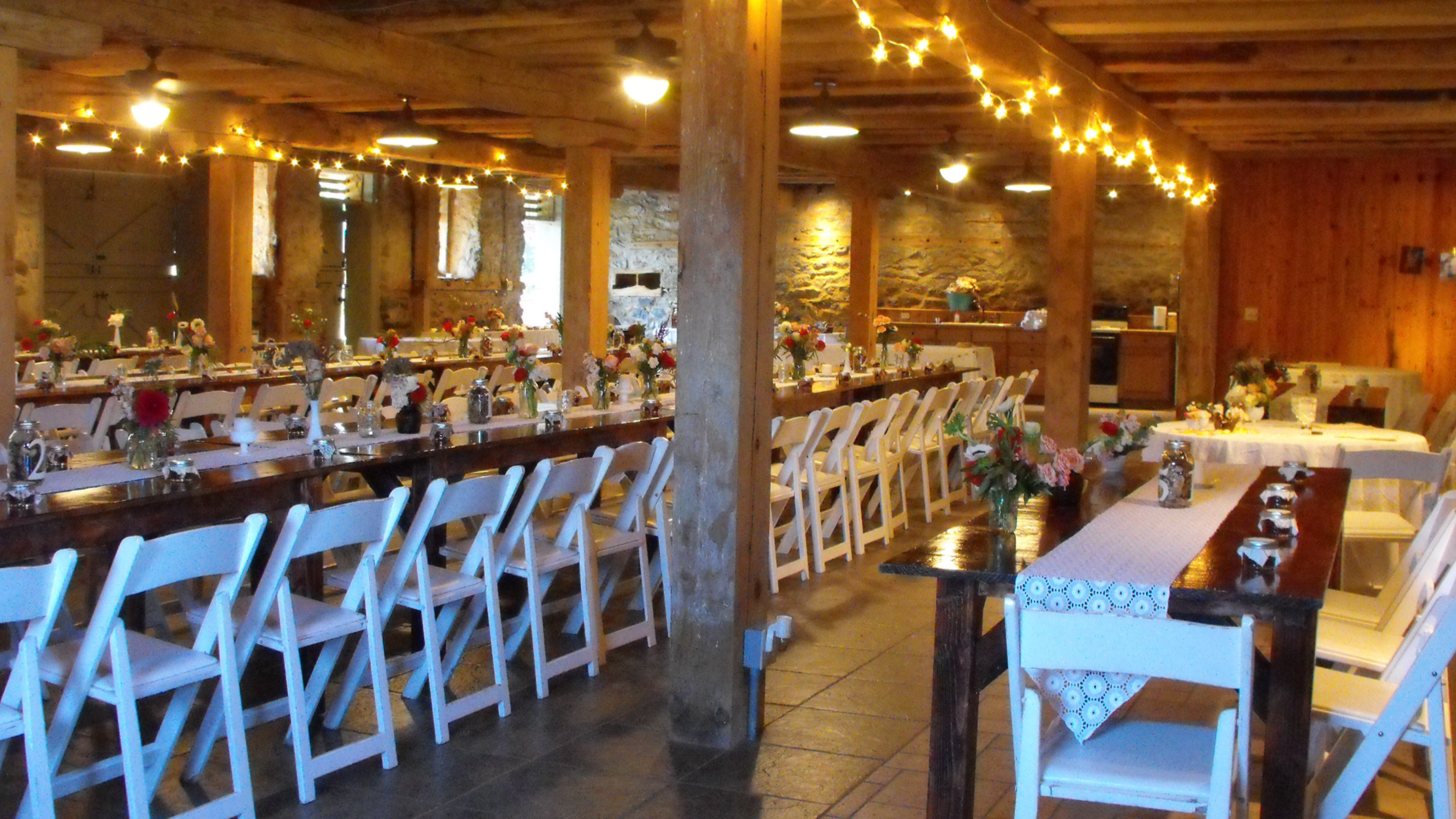 Indoor rustic party or event space with long tables decorated with flowers and candles, string lights hanging from ceiling, and wooden beams and walls.