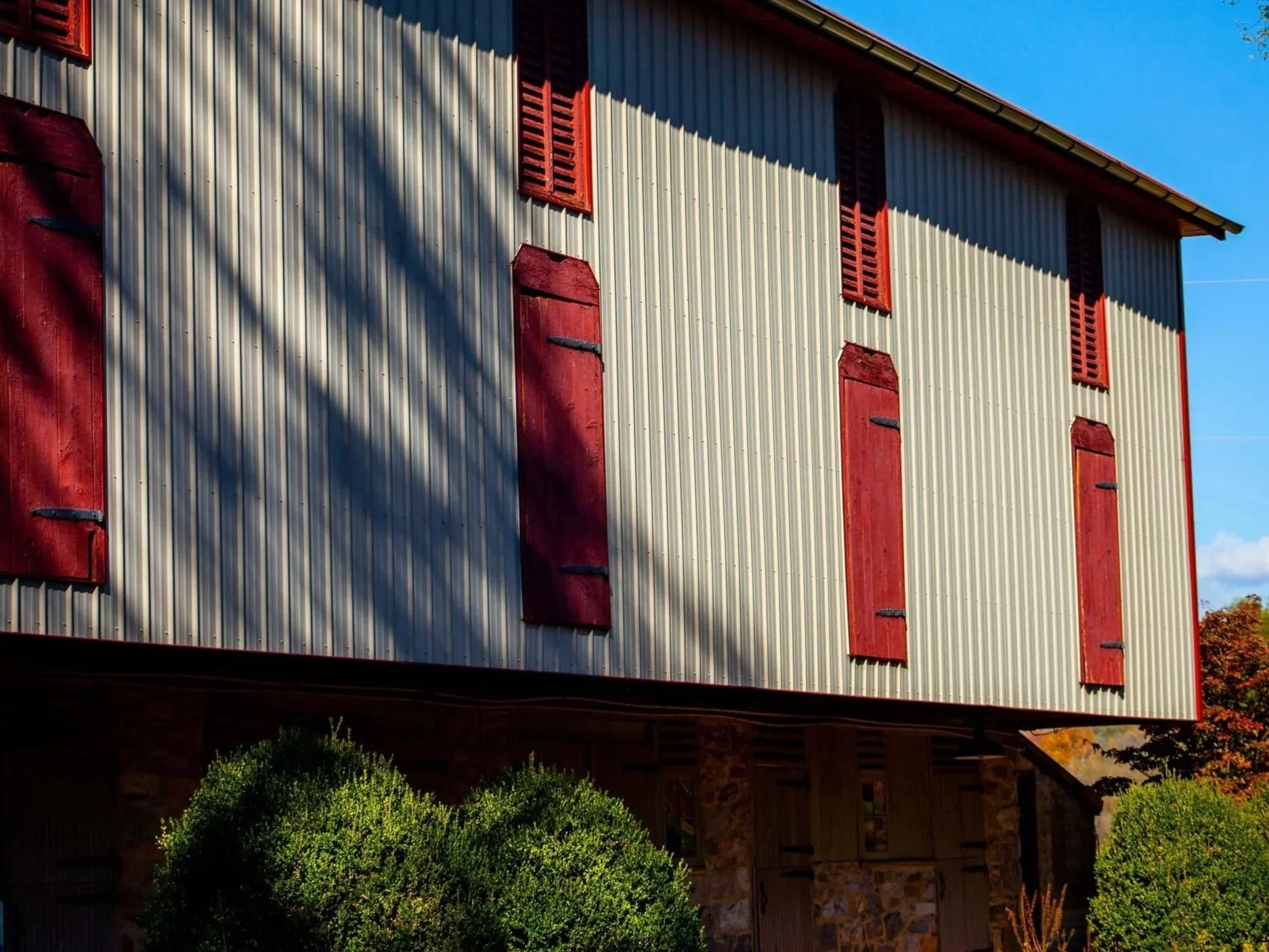 A metal building with red closed shutters on its windows, set against a blue sky with some clouds and trees in the background.