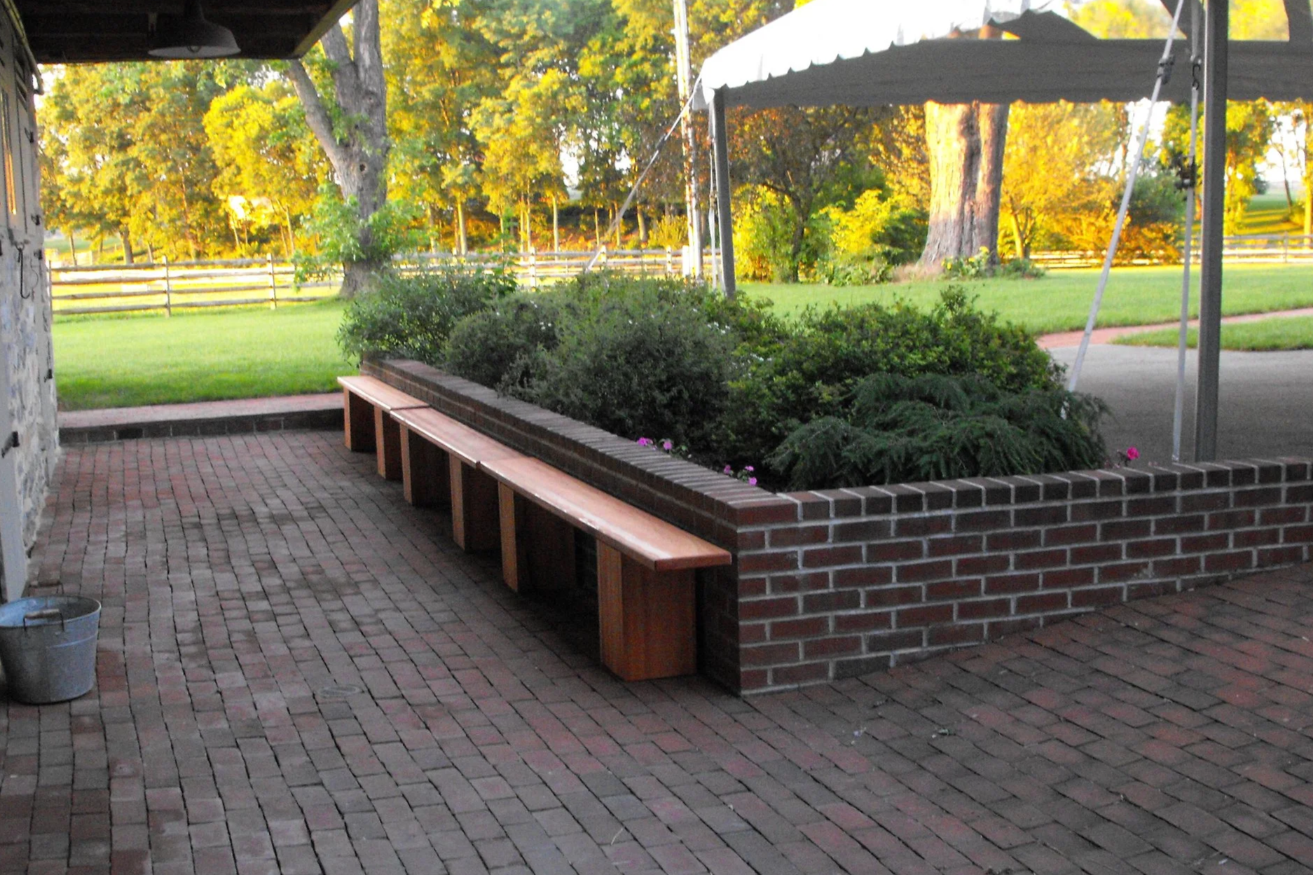 A brick-paved outdoor patio area with a long wooden bench, a flower bed with green shrubs, and a white canopy tent in the background. There is a grassy lawn and trees illuminated by sunlight.