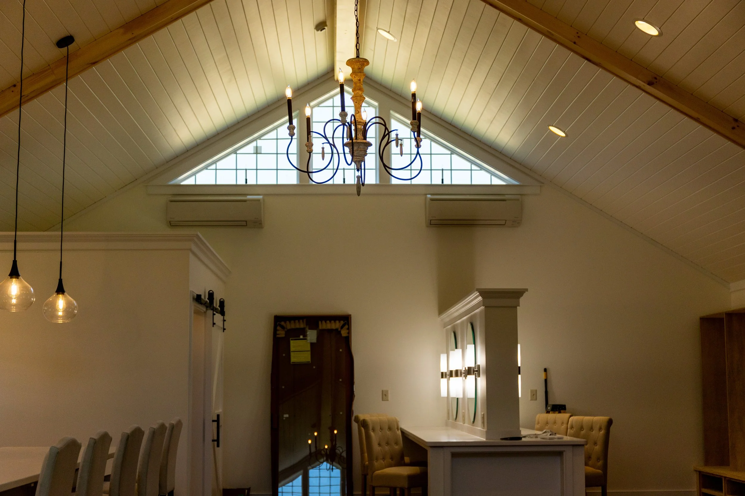 Interior of a room with a high, vaulted ceiling made of light wood panels, featuring two air conditioning units and a large window with decorative grid design. A chandelier with lit candles hangs from the ceiling, and a pair of hanging pendant lights
