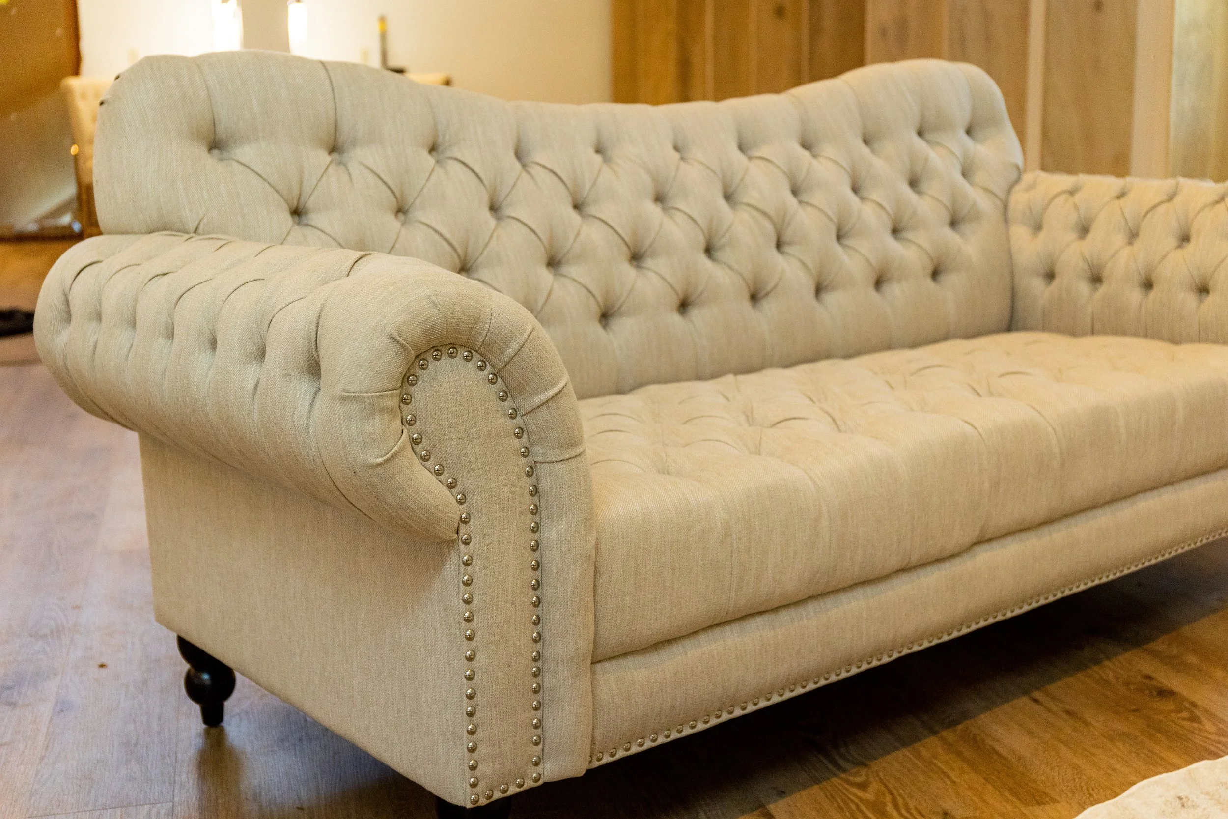 A beige upholstered sofa with tufted back and rolled arms, decorated with nailhead trim, sitting on wooden legs in a living room.