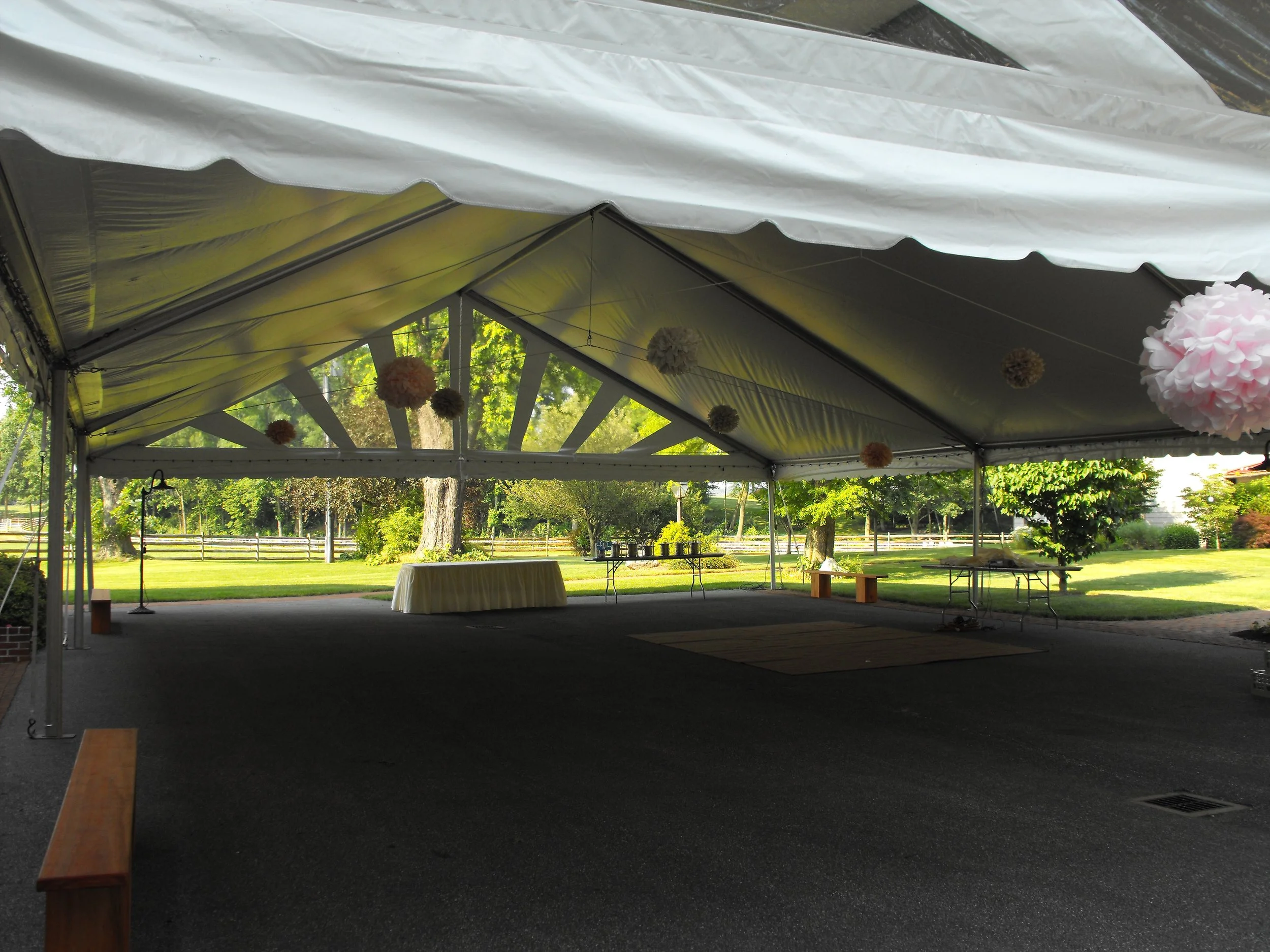 Empty outdoor event tent set up with hanging decorations, tables, and benches, with a green lawn and trees in the background.