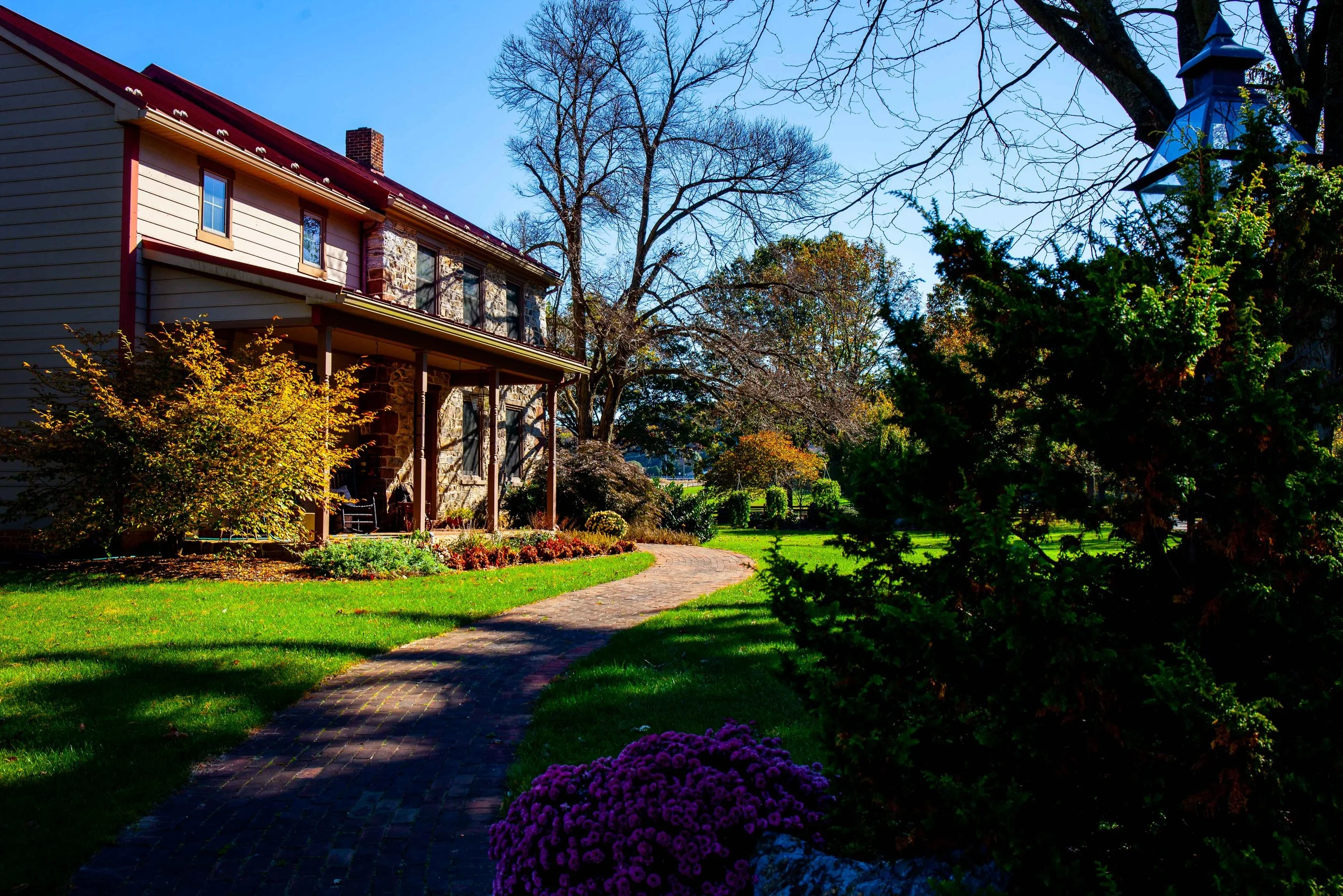 A house with a stone and wood exterior, a red roof, and a porch surrounded by a garden with colorful flowers, a winding brick pathway, trees, and a clear blue sky.