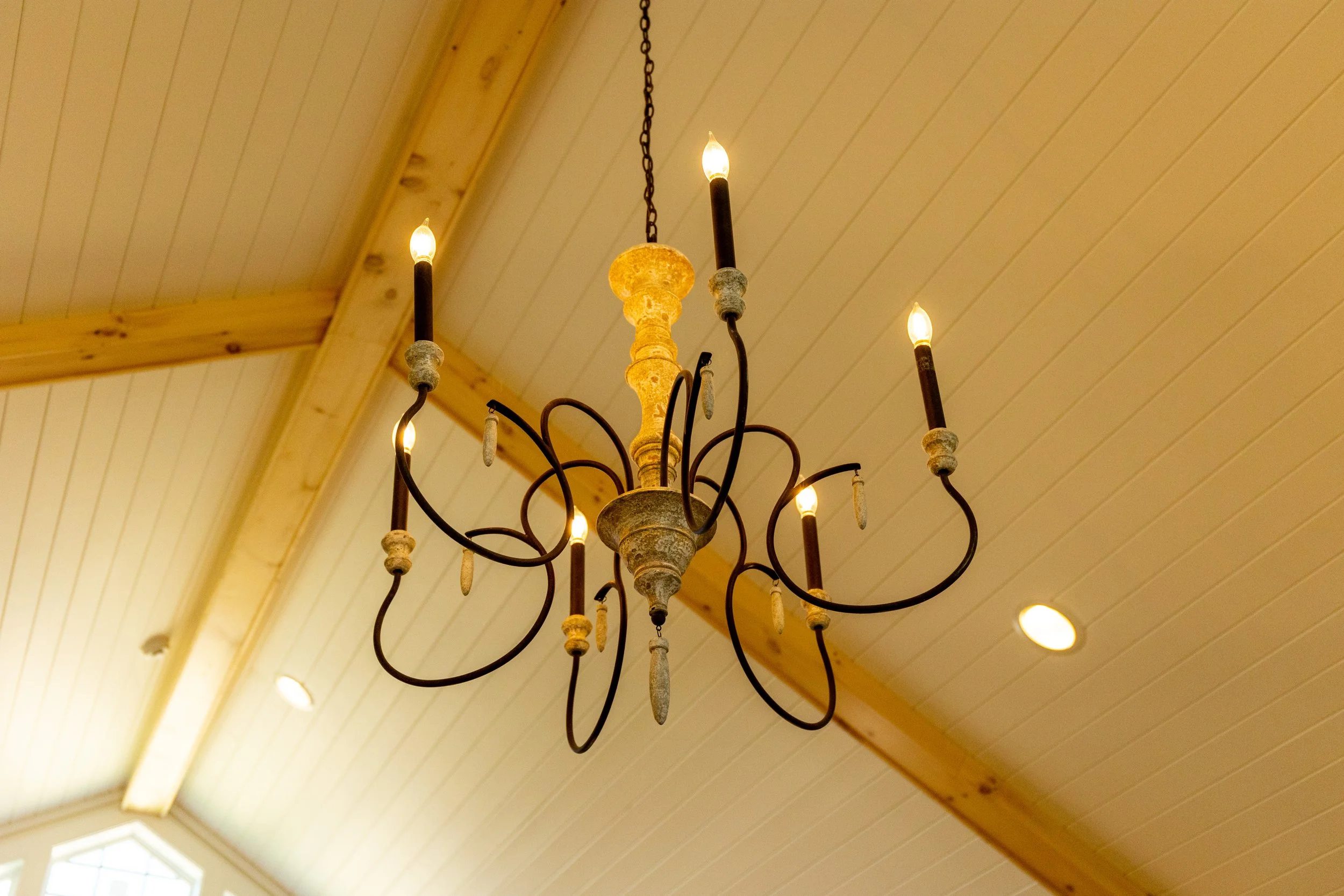 A chandelier with multiple arms and candle-shaped light bulbs hanging from a chain in a room with a wooden ceiling and beams.