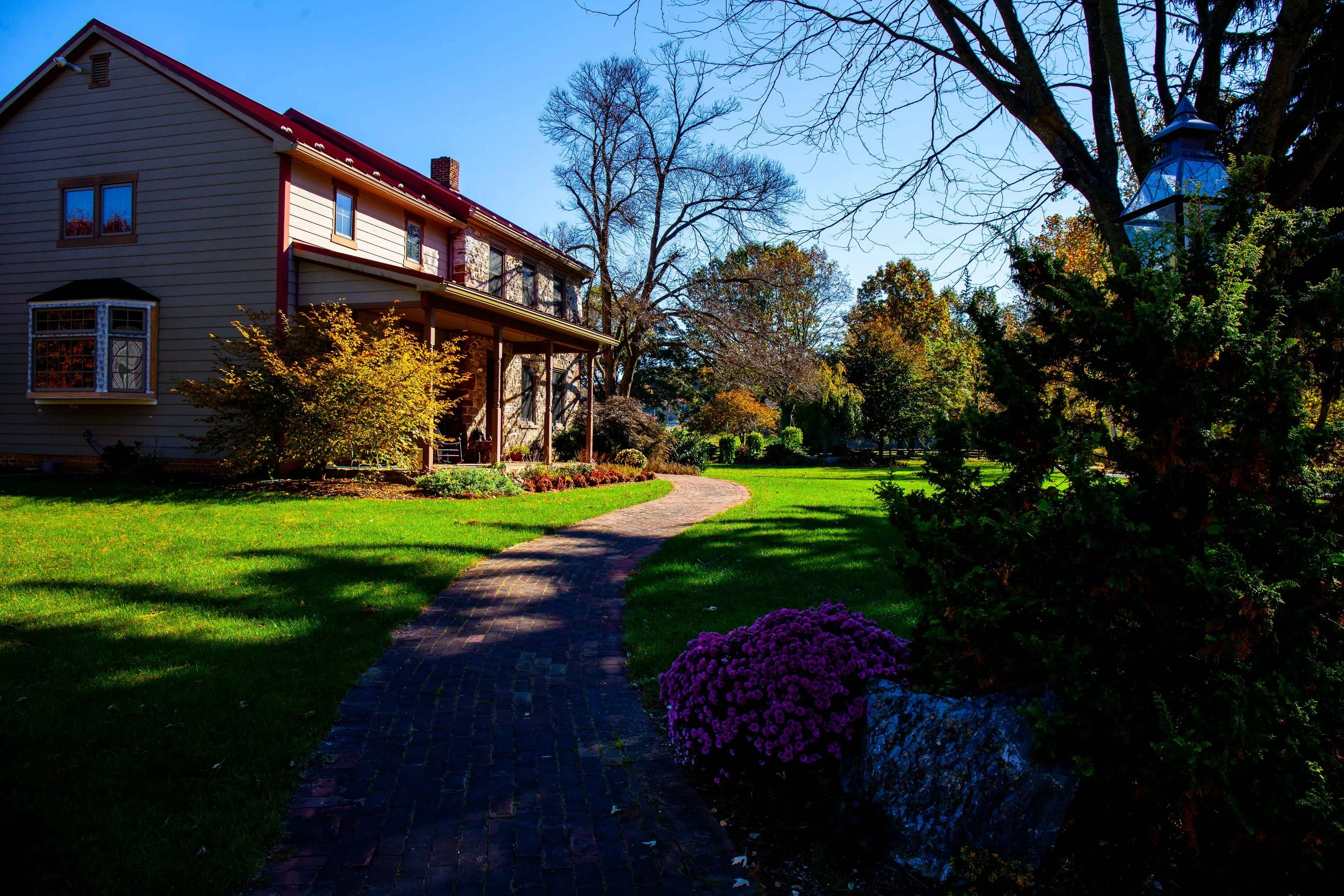 A house with a brick pathway leading to the front porch, surrounded by green grass and colorful trees, under a clear blue sky.