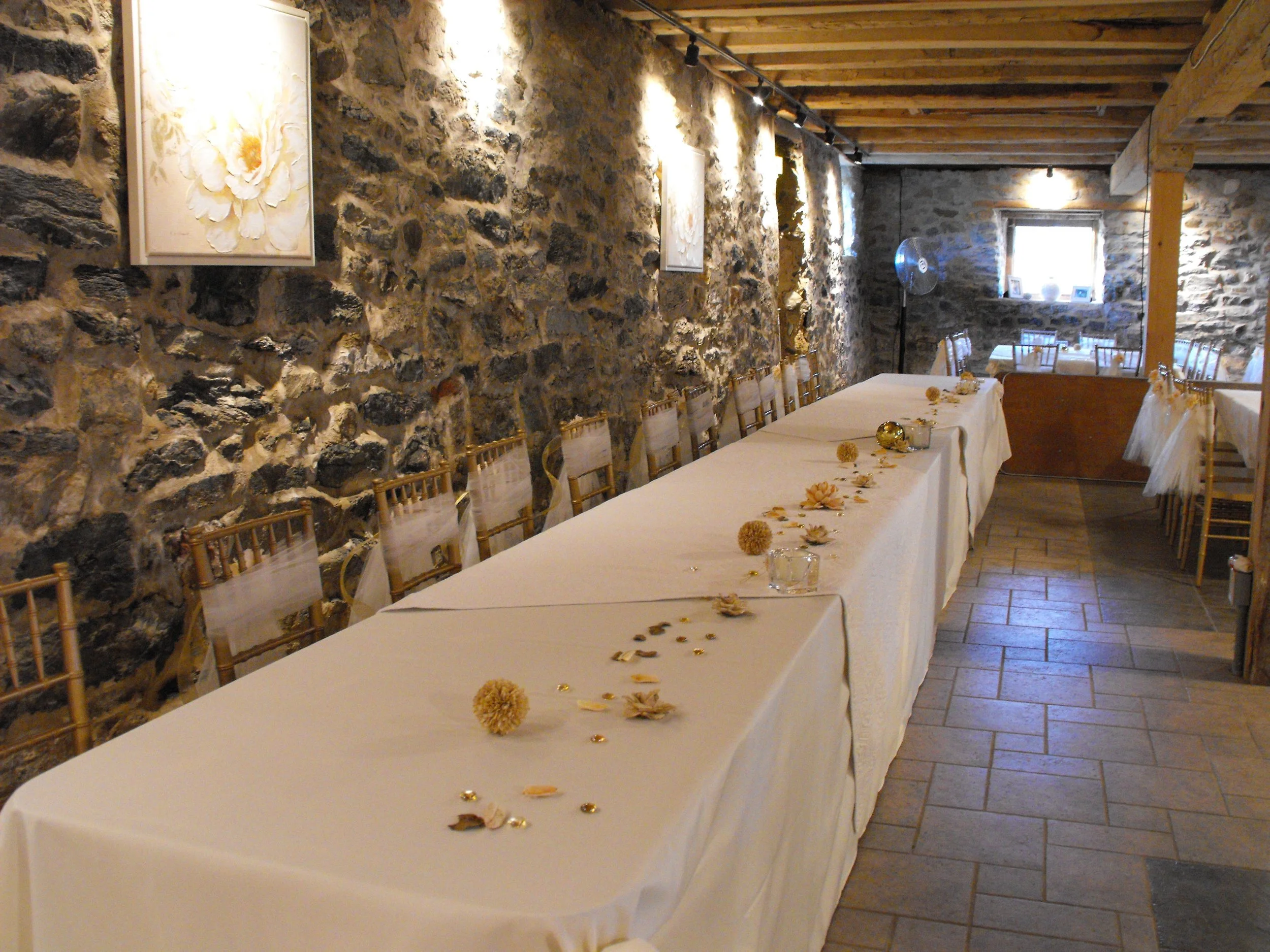 A long banquet table with a white tablecloth in a rustic stone-walled room decorated with flowers and candles, set for an event.