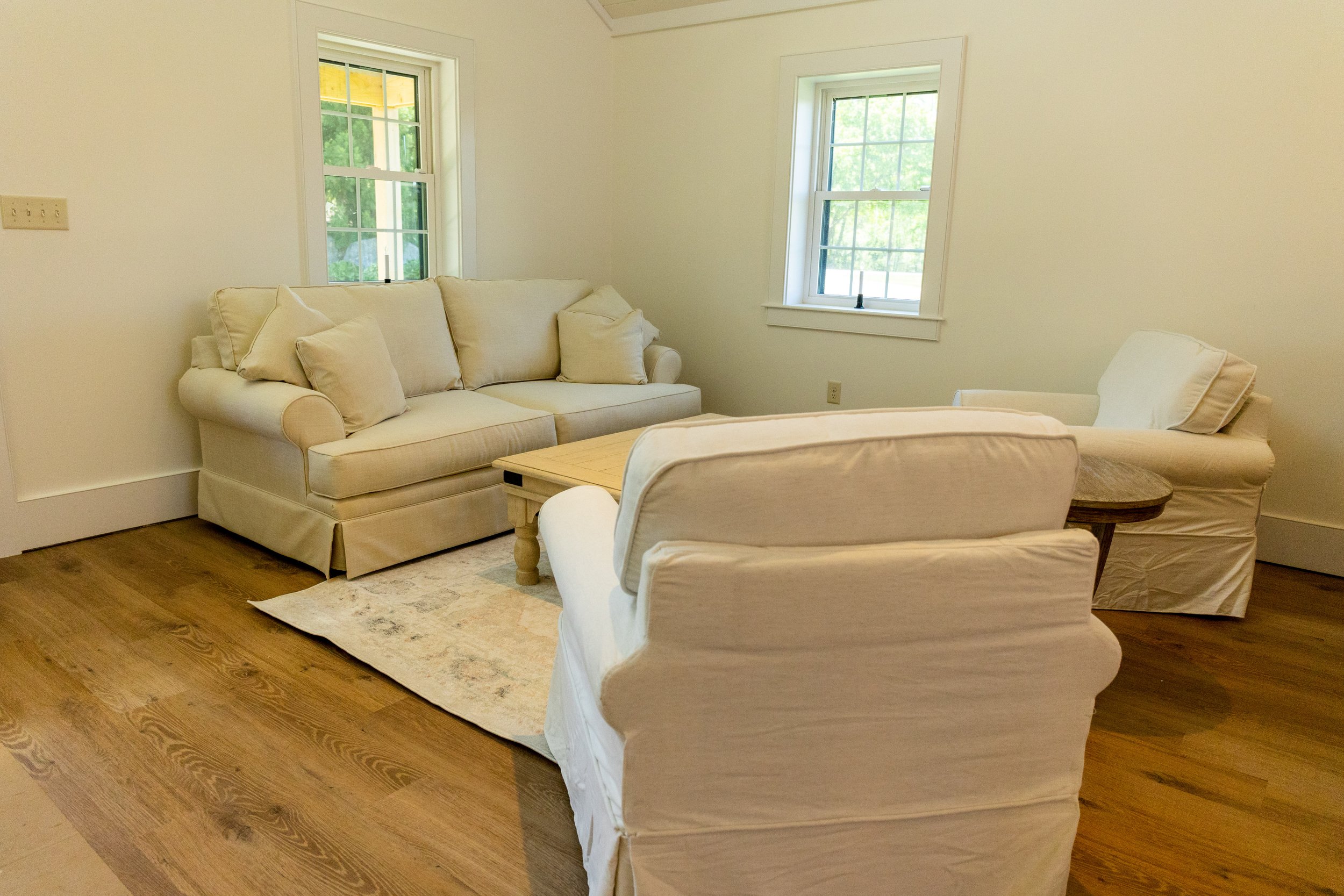 Living room with white sofa, two white armchairs, a wooden coffee table, a patterned area rug, and two windows showing greenery outside.