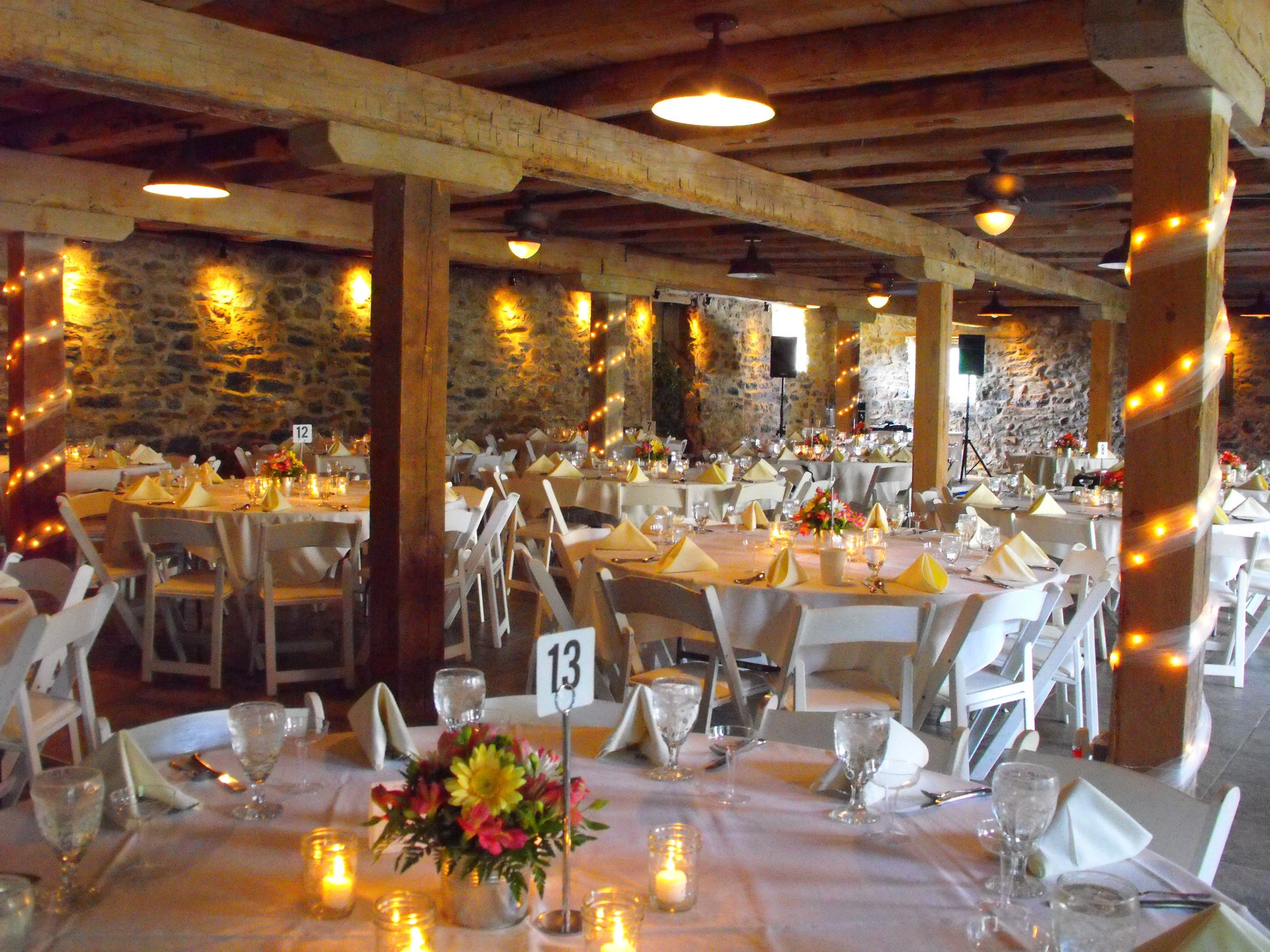 Indoor wedding reception with round tables covered in white tablecloths, decorated with flower centerpieces, candles, and neatly folded napkins, set against exposed stone walls with warm string lights and wooden beams on the ceiling.