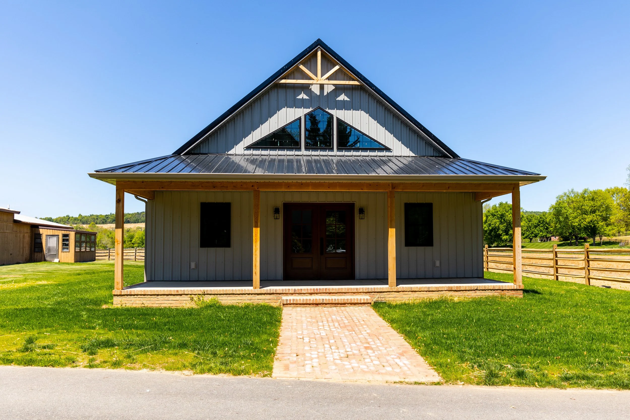 Front view of a modern farmhouse with metal roof, glass-paned double doors, on a green lawn with a brick sidewalk, wooden porch posts, and surrounded by a wooden fence, under a clear blue sky.