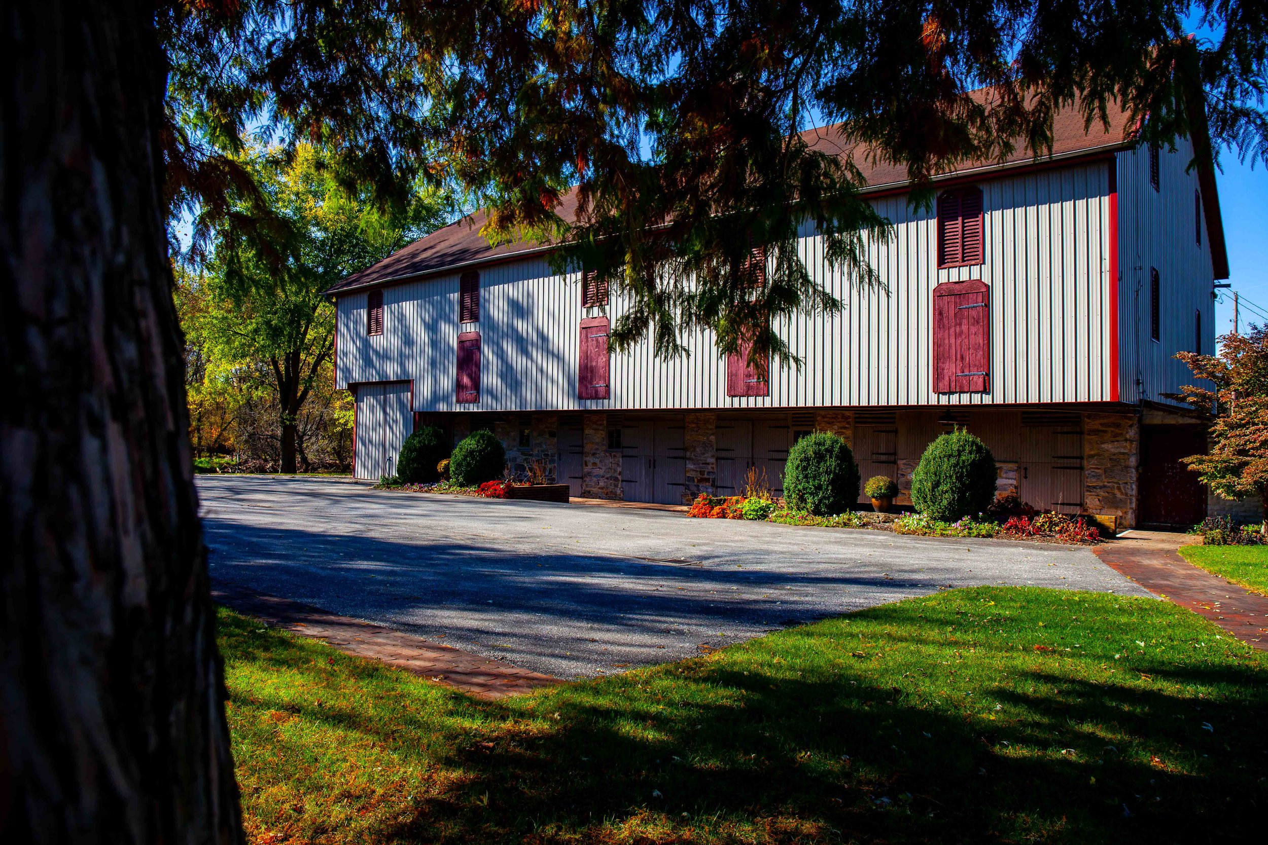 A large, two-story barn with metal siding and wooden shutters, surrounded by trees with colorful autumn leaves, and a driveway in front.