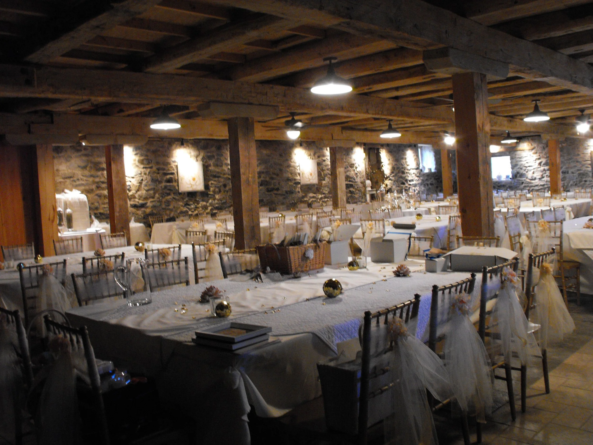 Long banquet table decorated with white tablecloths, gold ornaments, and tulle bows on chairs in a rustic stone-walled event space with wooden beams and hanging lights.
