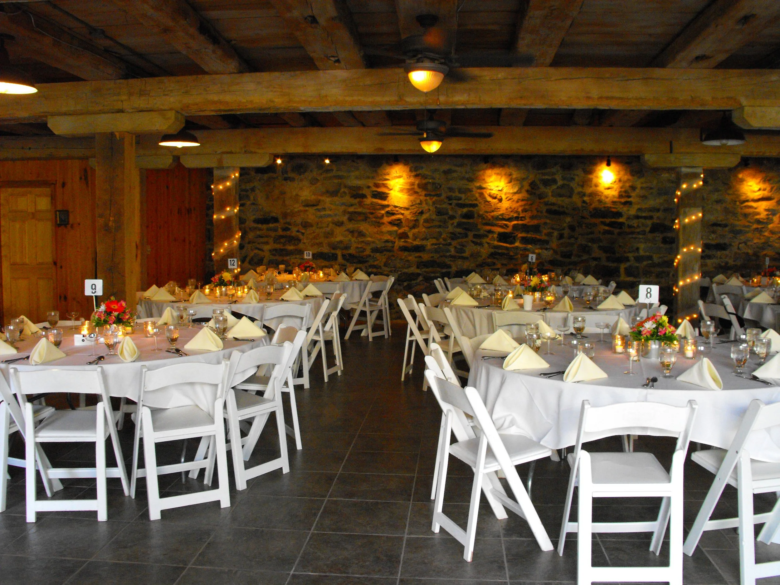 A banquet hall decorated for a formal event with multiple round tables covered in white tablecloths, each set with napkins, glassware, and flowers, against a background of a stone wall and wooden beams.