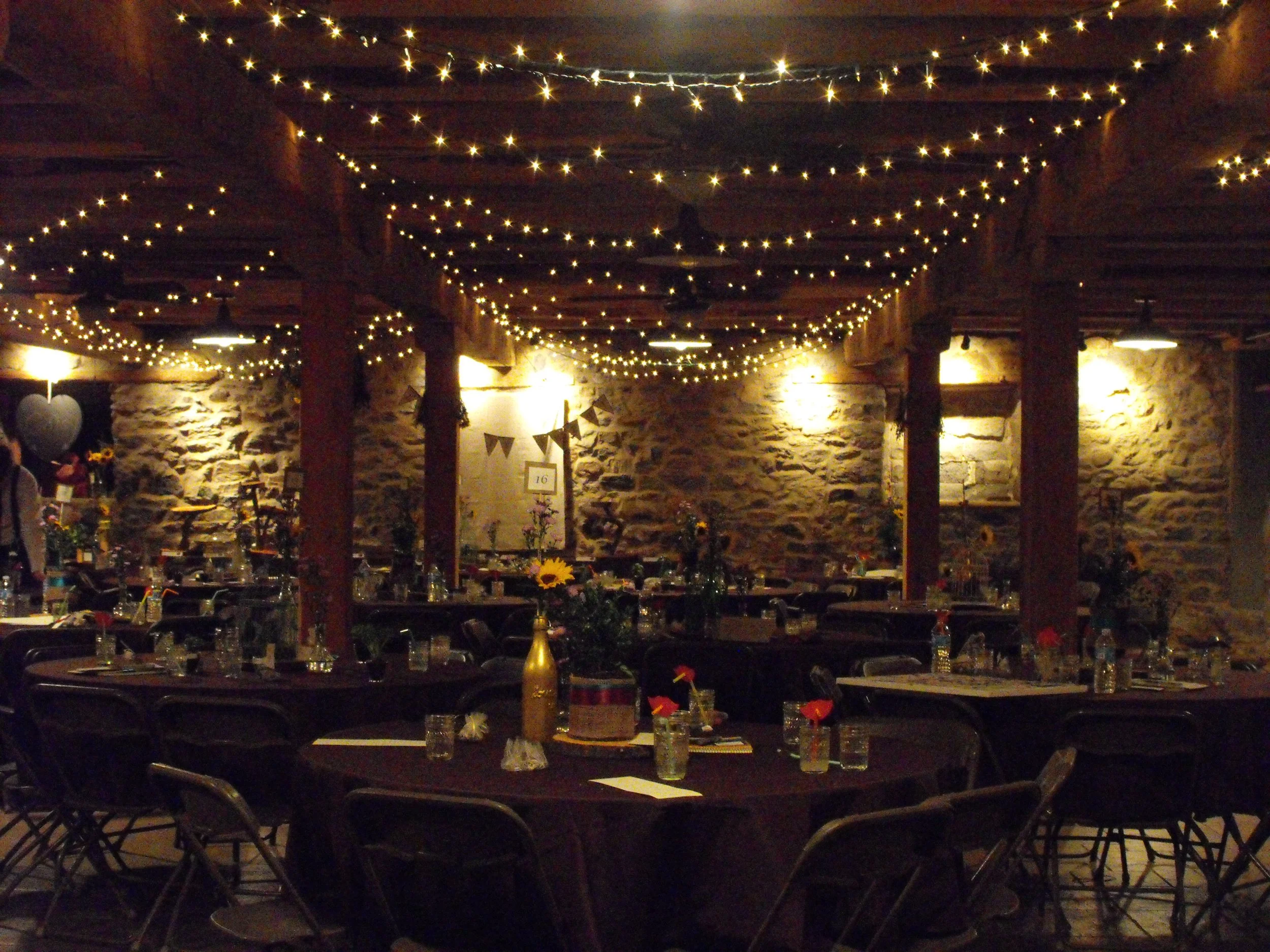 Indoor event space decorated with string lights hanging from the ceiling, tables set with glasses, bottles, and tableware, and stone walls in the background.